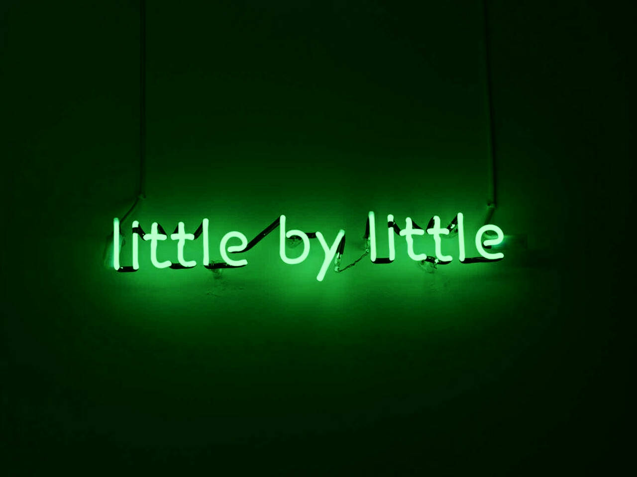 lights 桌布 壁紙 — 一個發光的綠色霓虹燈牌，上面寫著 little by little，掛在黑暗的牆上，投射出柔和的环境光。