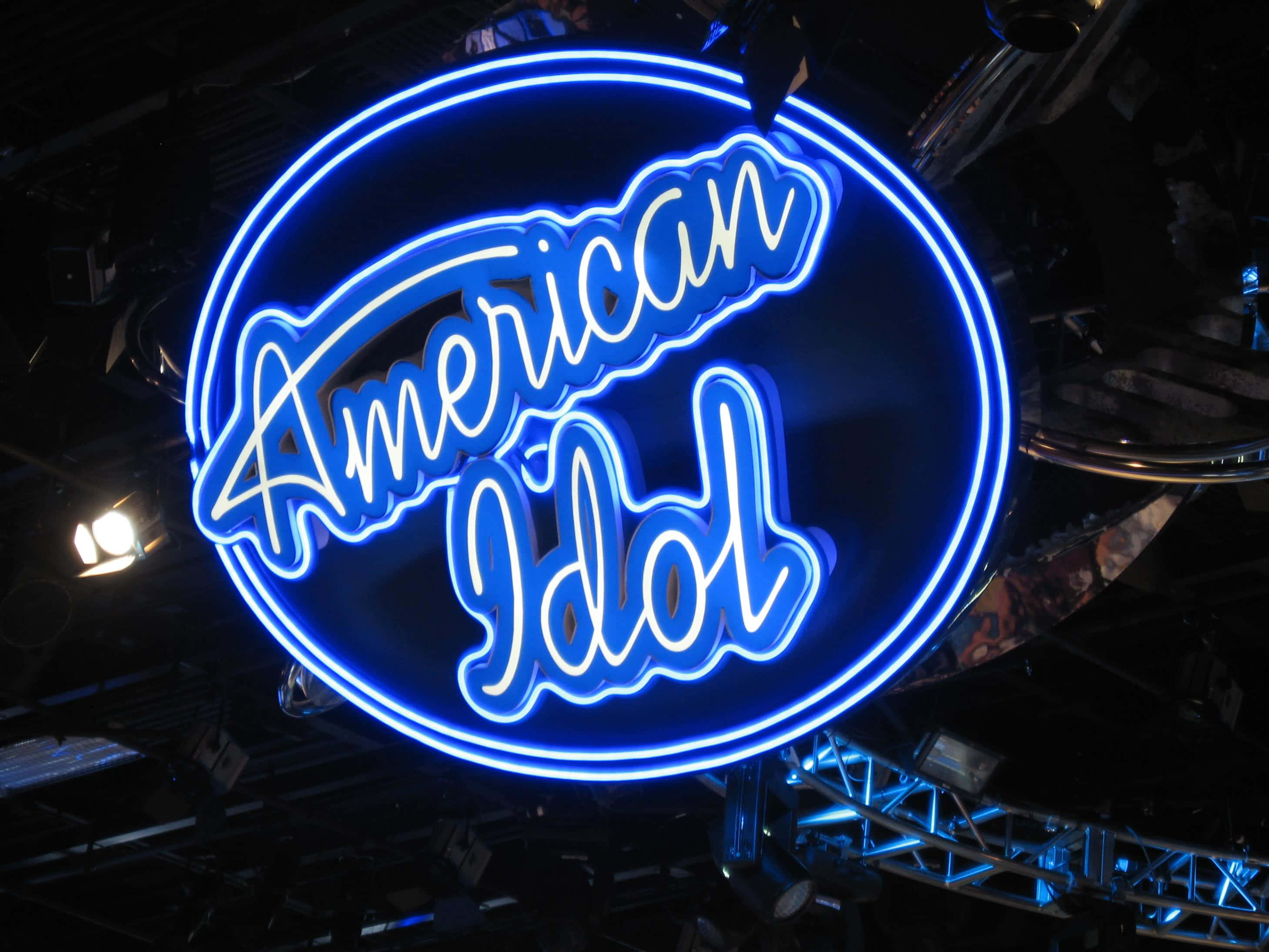 american idol 桌布 壁紙 — 一個發光的藍色American Idol霓虹燈牌在黑暗的舞台背景上明亮地閃耀，並可見懸掛裝置。
