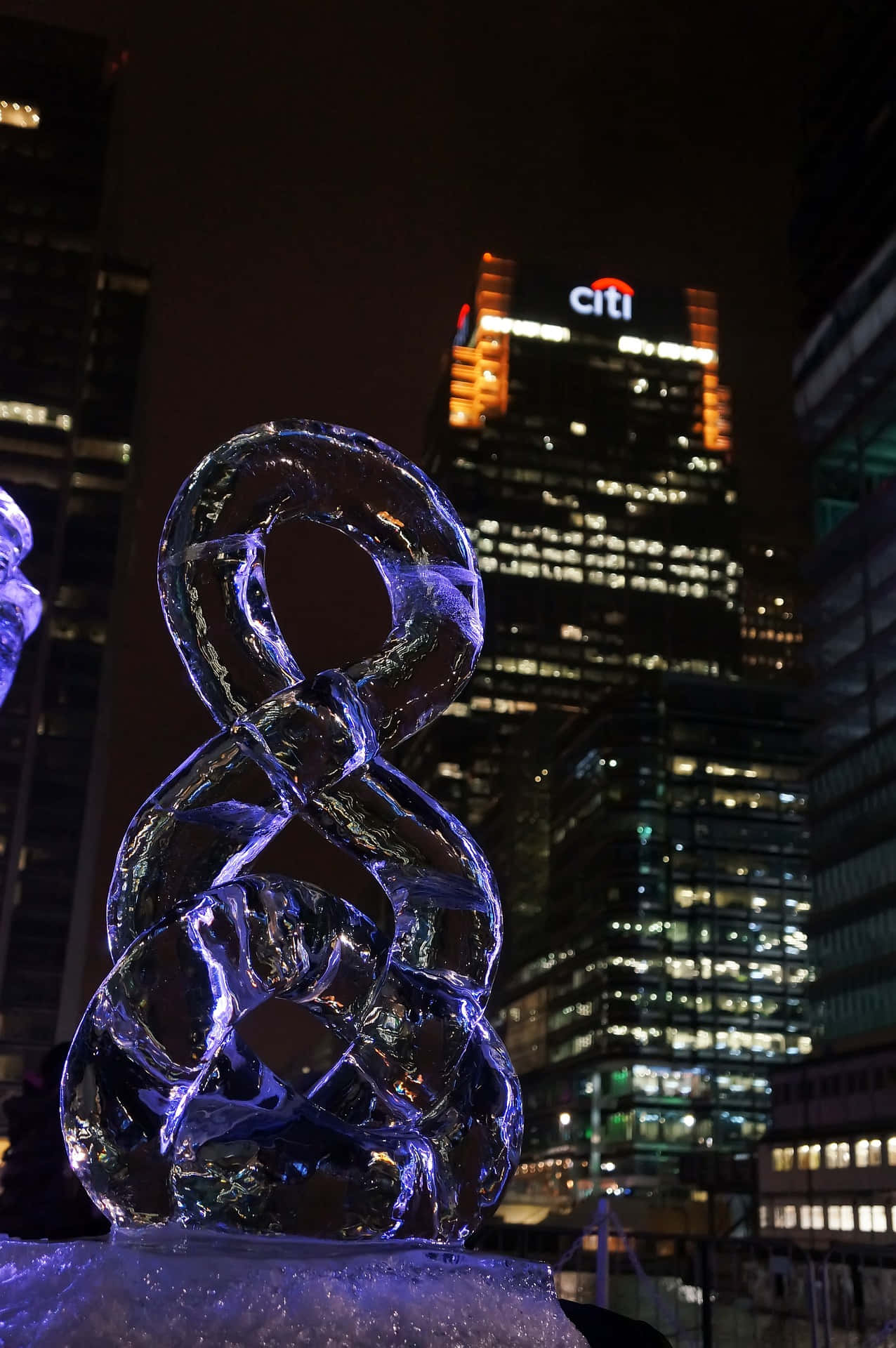 Ice Sculpture 桌布 壁紙 — 一個發光的藍色冰雕，帶有複雜的環圈，夜晚矗立在燈火通明的花旗摩天大樓前。