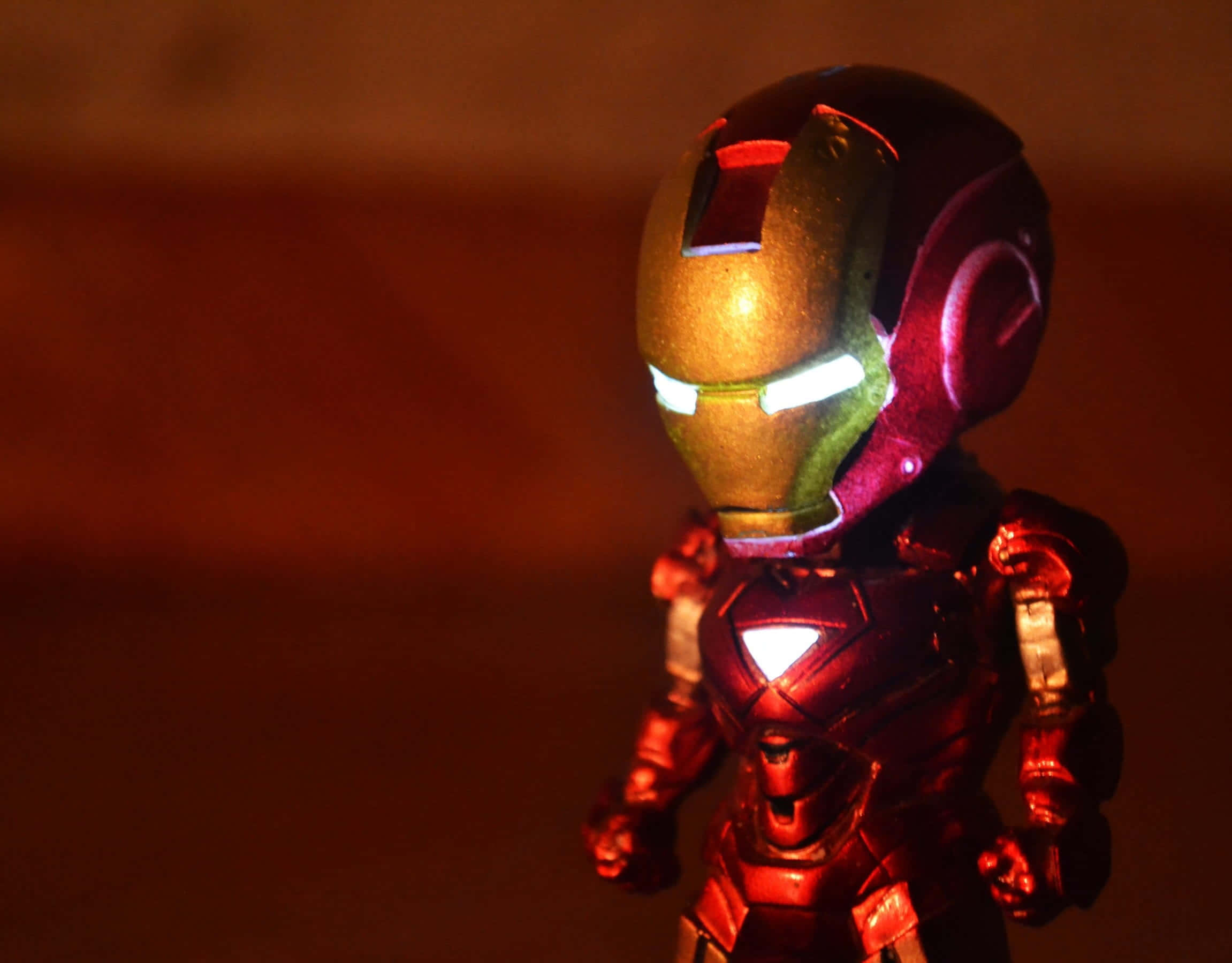 Iron Man Action Figures 桌布 壁紙 — 一個發光的鐵甲奇俠人偶站在昏暗燈光下，眼睛和胸部弧形反應器發光，背景溫暖模糊。