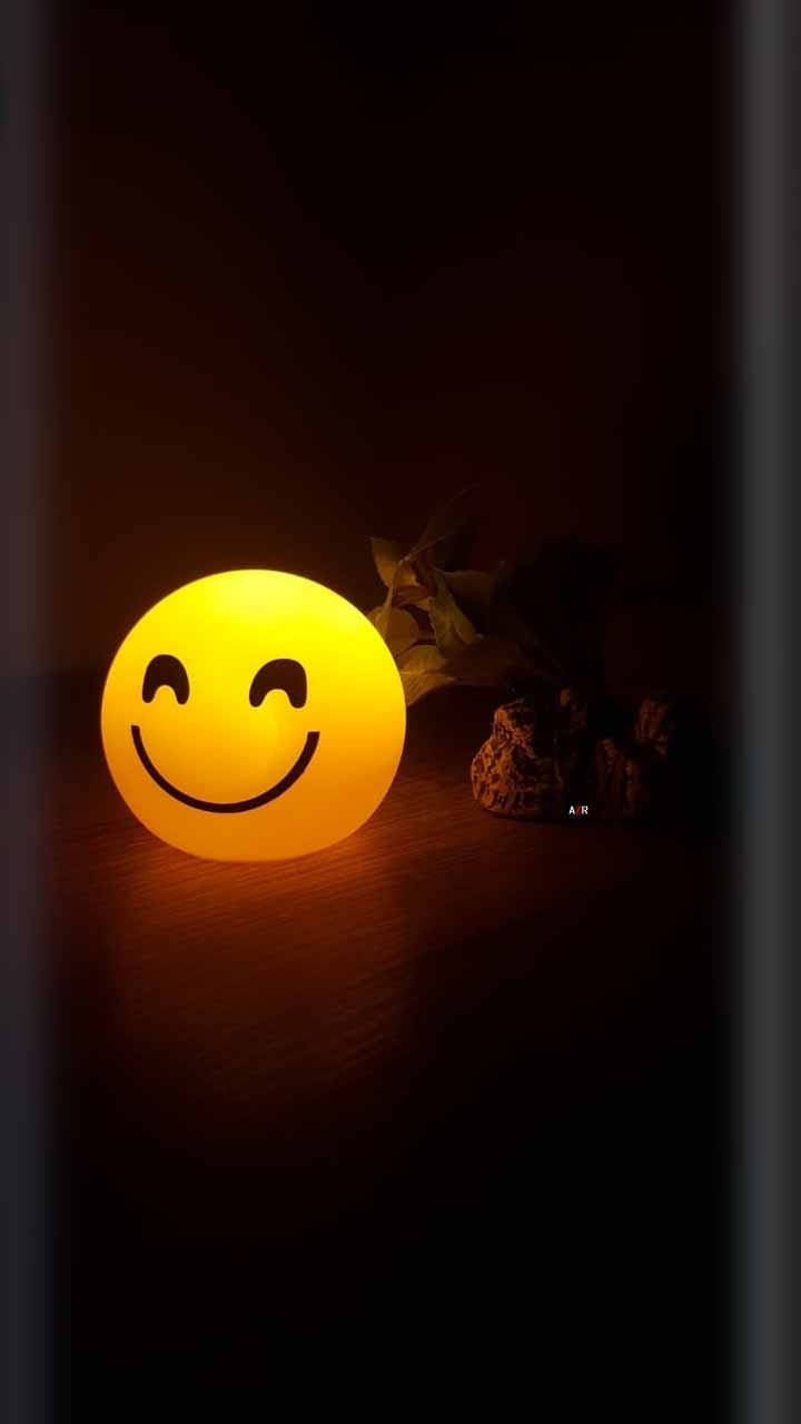 笑哈哈表情 桌布 壁紙 — 一個發光的黃色笑臉燈在黑暗的房間中，將溫暖的光線投射在木質表面和旁邊的乾葉子上。
