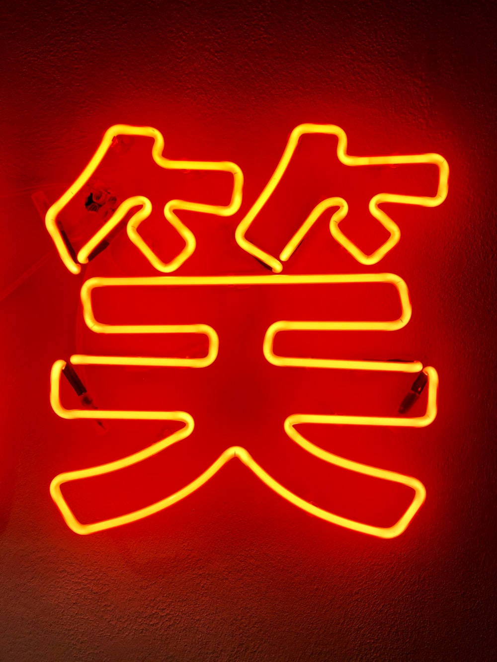 Laugh 桌布 壁紙 — 一個發光的黃色霓虹燈牌，形狀像中文的「笑」字，在紋理牆壁上投射出溫暖的紅光。