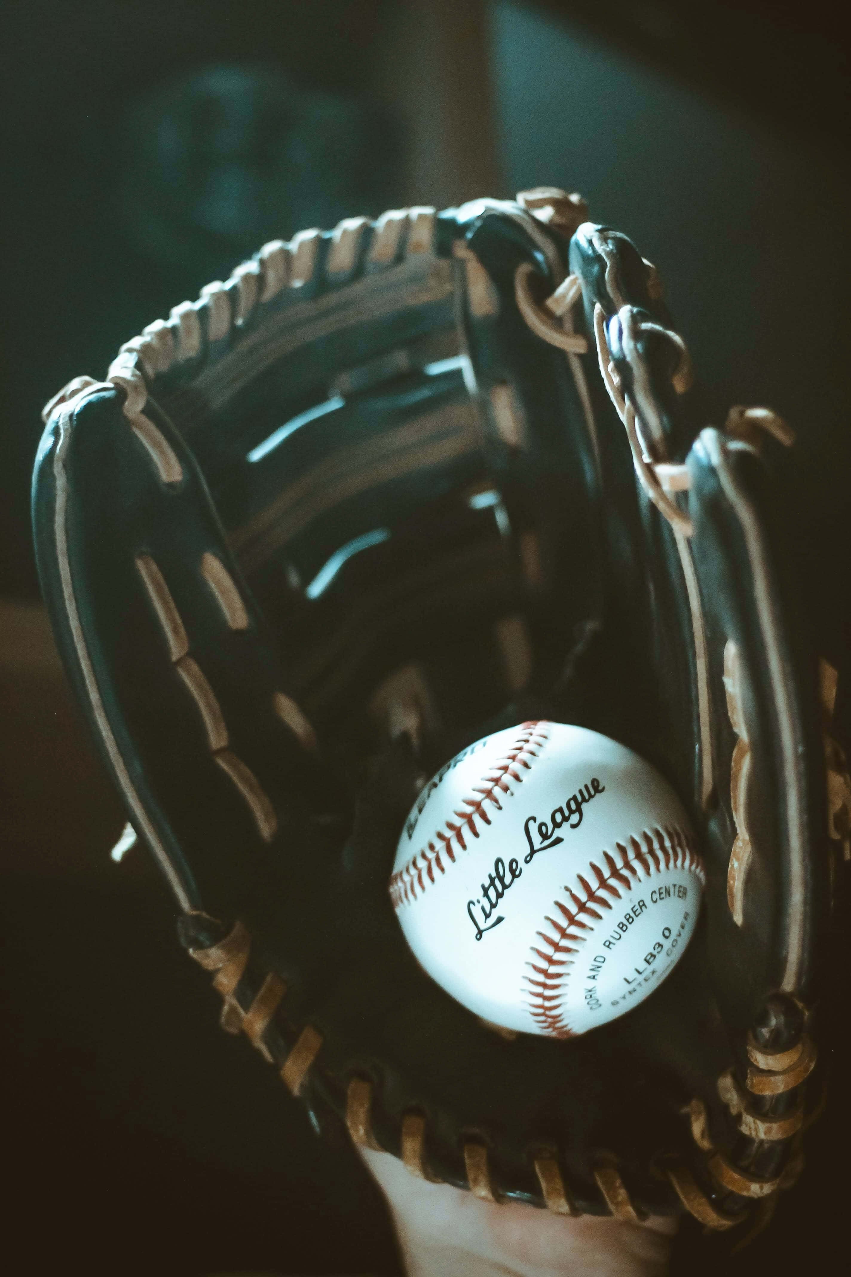 Baseball Gloves 桌布 壁紙 — 一個白色Little League棒球的特寫，放在一個深色皮革手套中，縫線清晰可見，有一隻手握住它。
