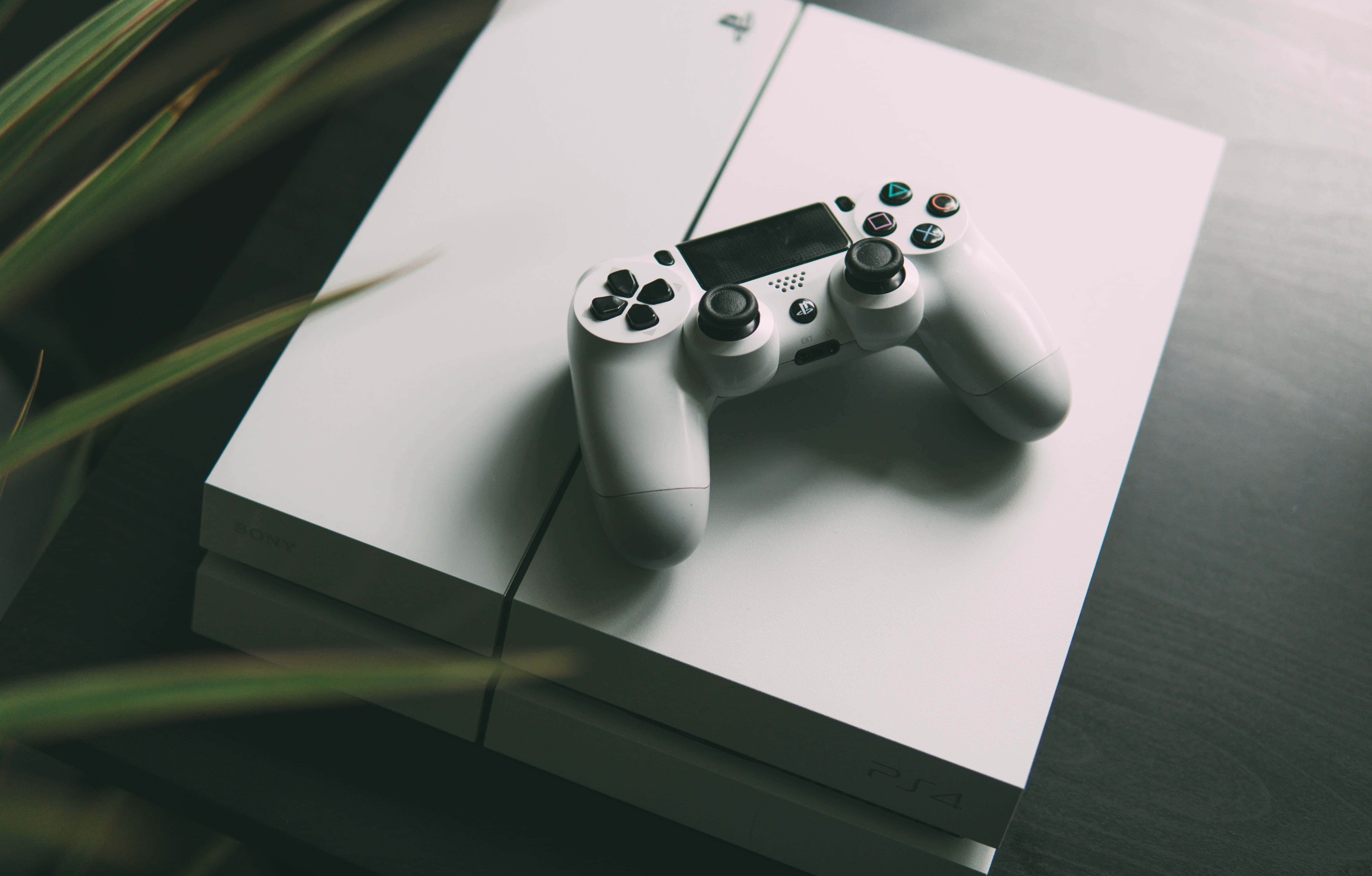 Gaming Consoles 桌布 壁紙 — 一個白色PlayStation 4手掣放在主機上，柔和自然光線，前景有模糊的綠色植物葉子。
