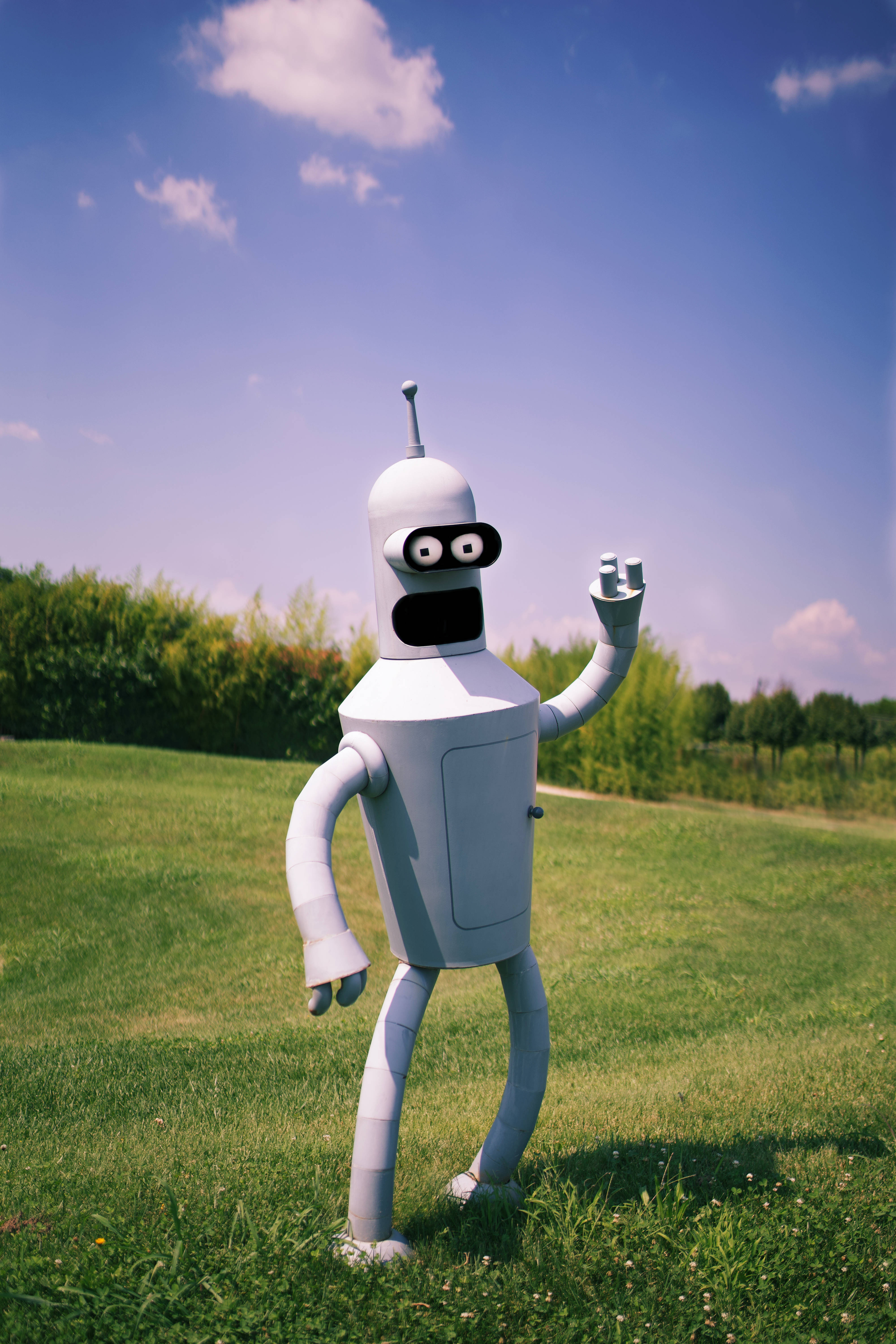 Futurama Realistic Bender 壁紙 — 一個白色卡通機械人，大眼睛在陽光普照的綠色田野上，藍天白雲下開心地揮手。
