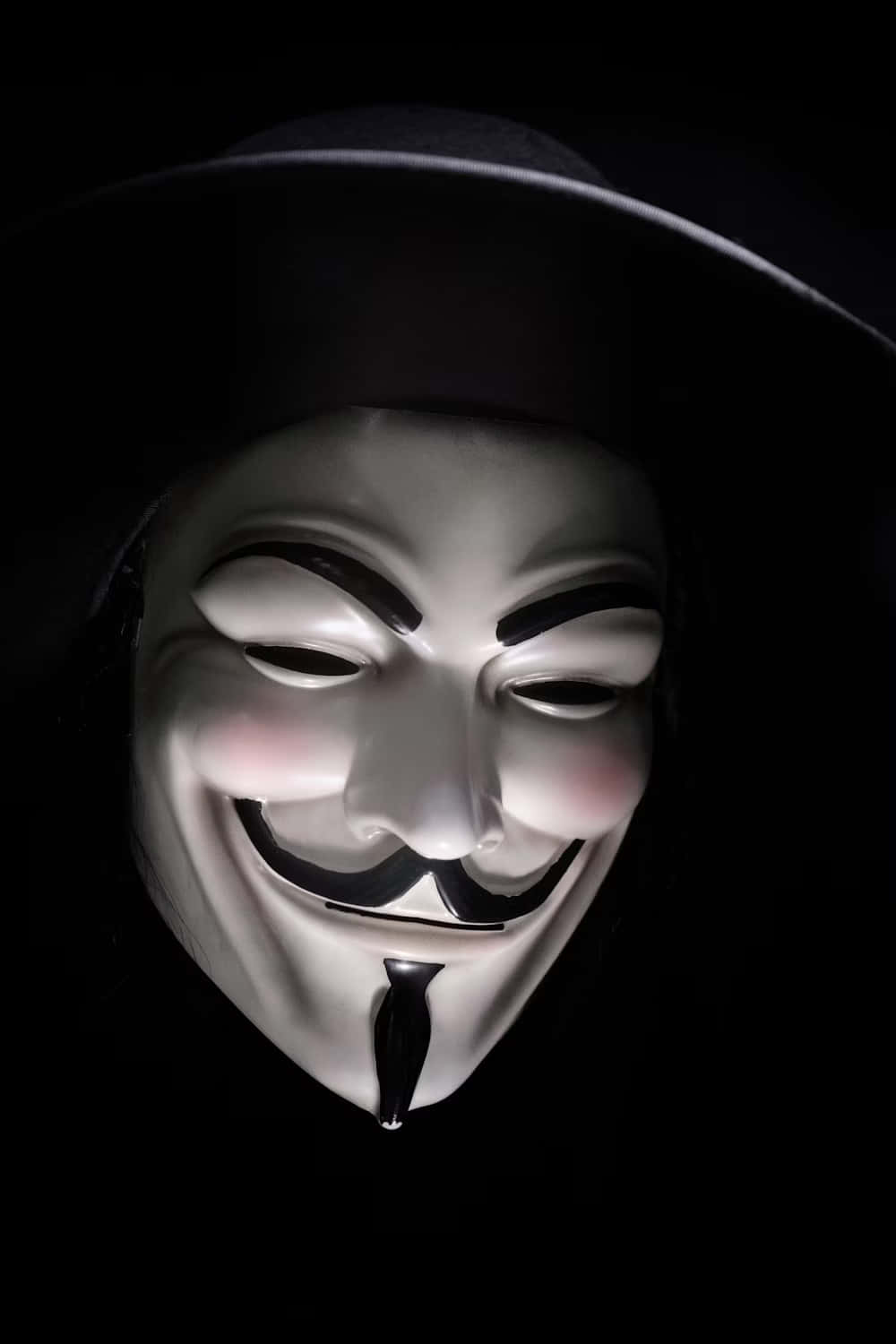  壁紙 — 一個白色嘅 Guy Fawkes 面具，帶住一個大笑容同黑色帽，喺漆黑背景前被照亮。