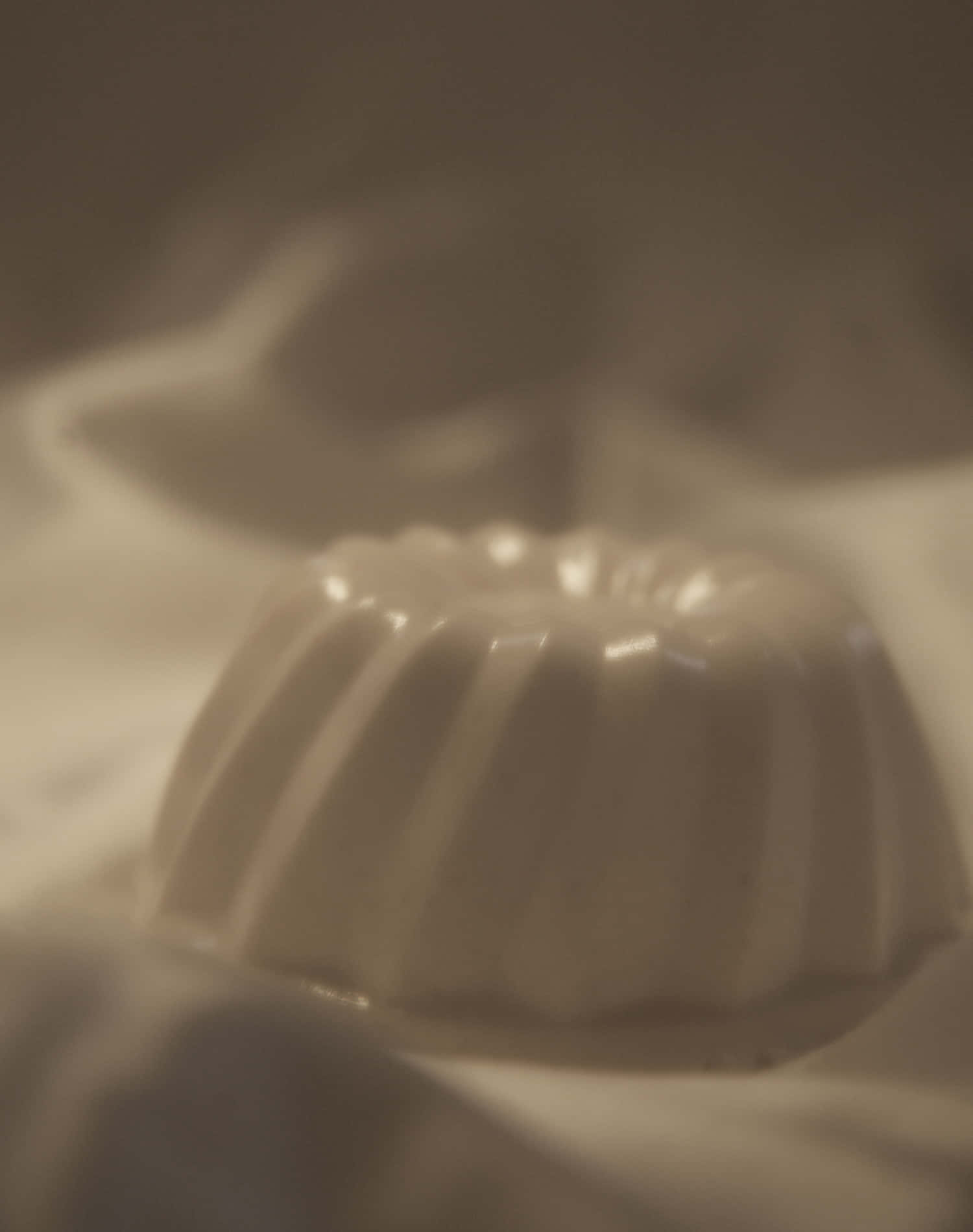 Blancmange 桌布 壁紙 — 一個白色帶凹槽的甜品模具特寫，放置在柔軟布料上，周圍有溫暖的環境光線。
