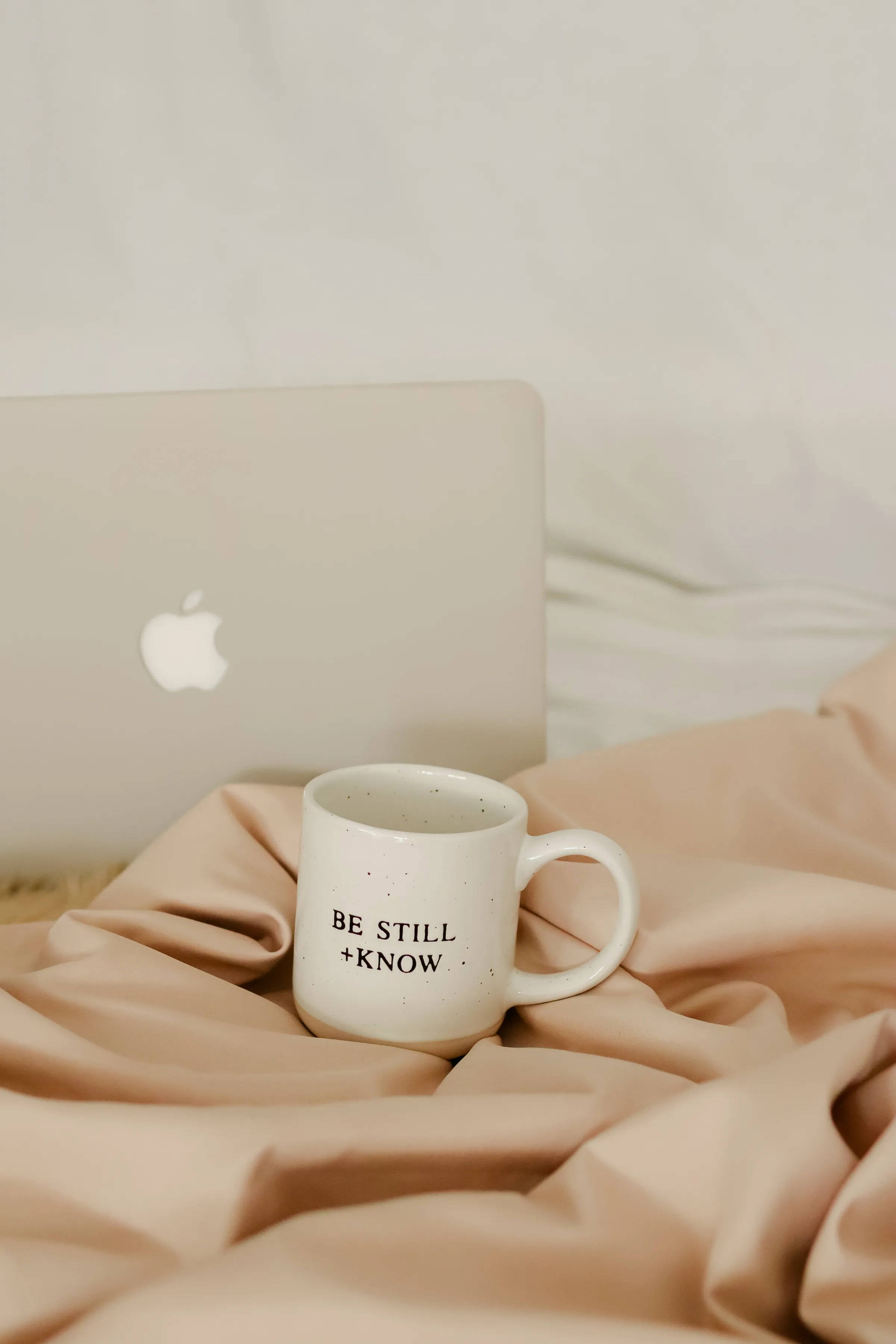 Coffee Aesthetic 桌布 壁紙 — 一個白色斑點馬克杯，上面寫著「BE STILL + KNOW」，放在床上柔軟的米色布料上，旁邊有一部銀色手提電腦。