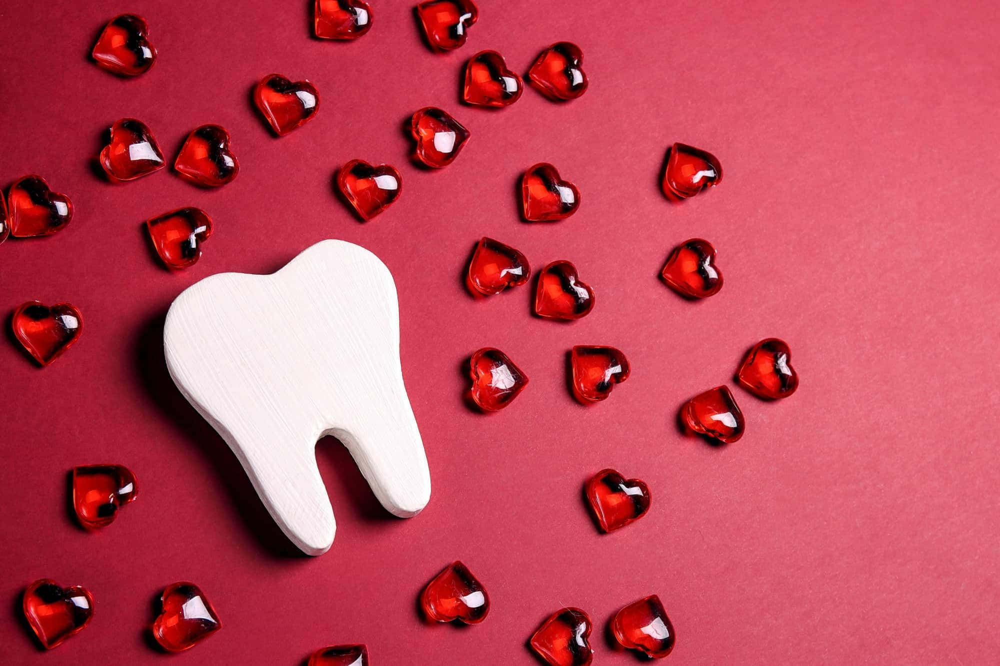 Tooth 桌布 壁紙 — 一個白色木製牙齒模型，周圍環繞著閃亮的紅色心形糖果，背景為深紅色。