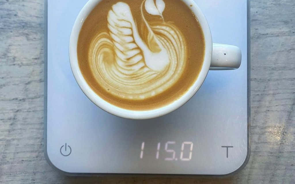 Milk And Coffee 桌布 壁紙 — 一個白色杯子帶有天鵝拉花藝術，放在一個顯示115.0克的數字秤上，置於木質表面。