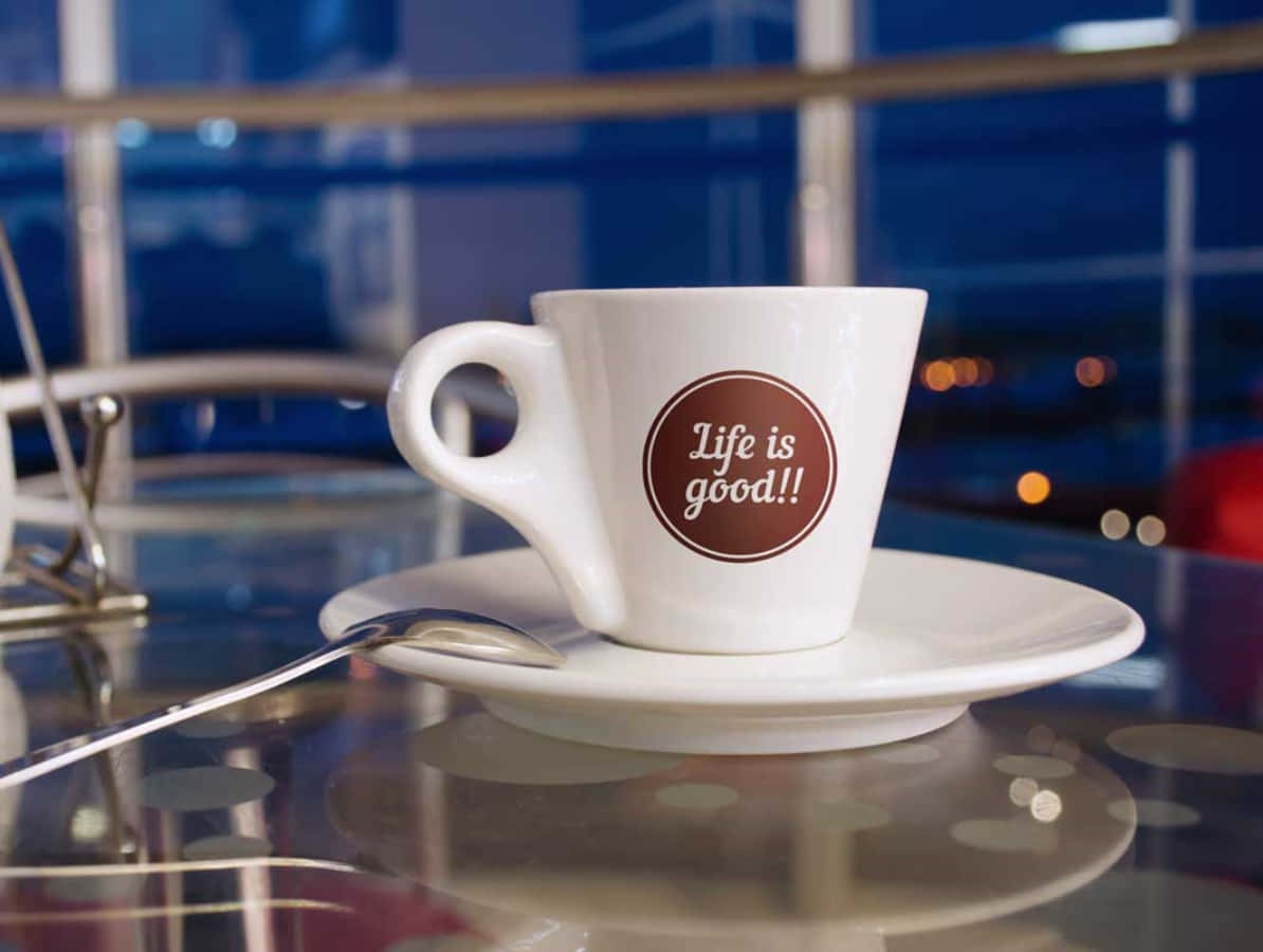 Coffee Cup 桌布 壁紙 — 一個白色濃縮咖啡杯上寫著「Life is good!!」，放在碟子上，置於反光玻璃桌上，背景是模糊的城市燈光。