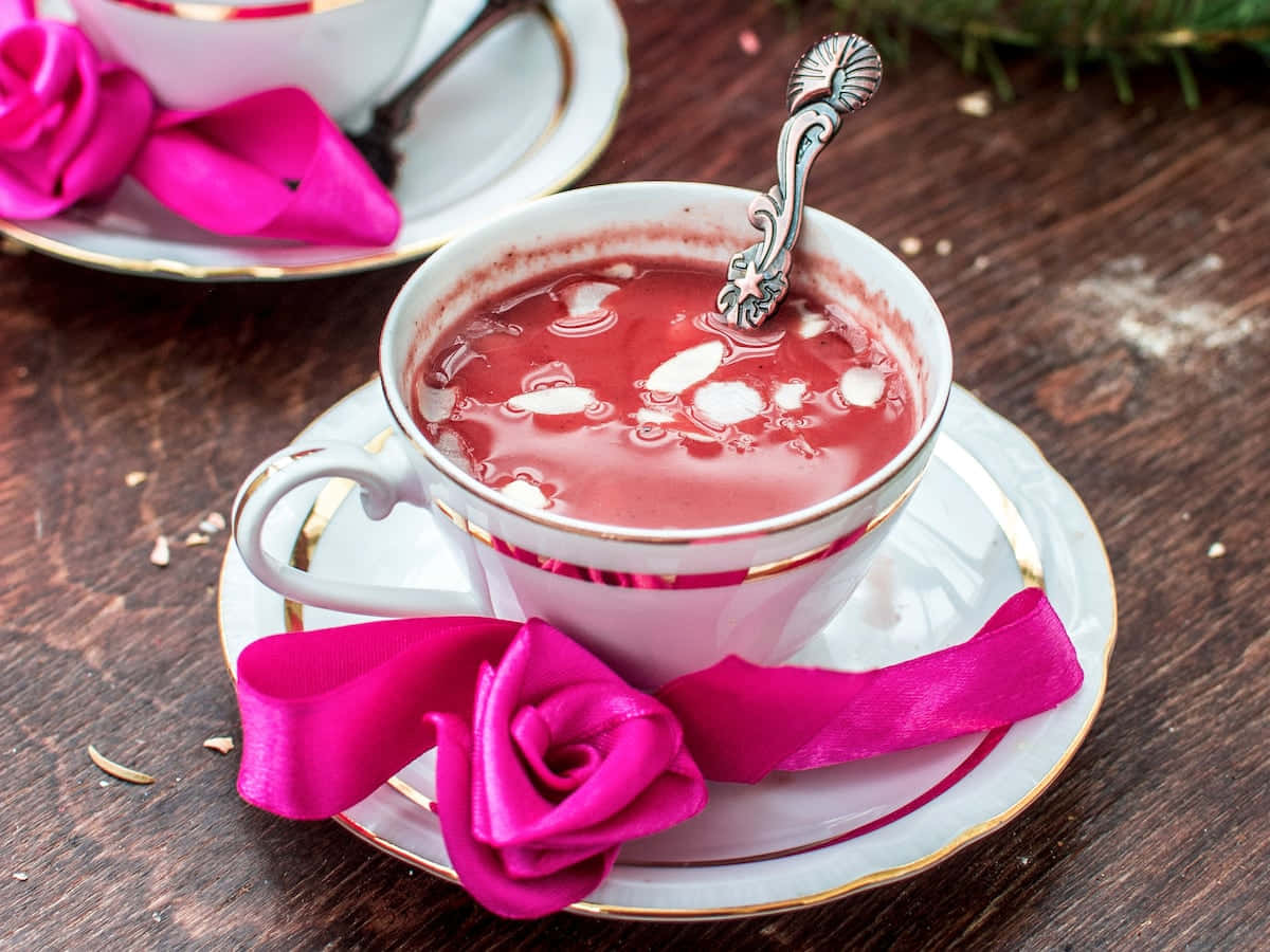 Pink Tea 桌布 壁紙 — 一個白色瓷杯裝滿充滿活力的粉紅色熱飲和奶油，放在一個飾有紫紅色緞帶玫瑰蝴蝶結的碟子上，置於鄉村風木桌上。