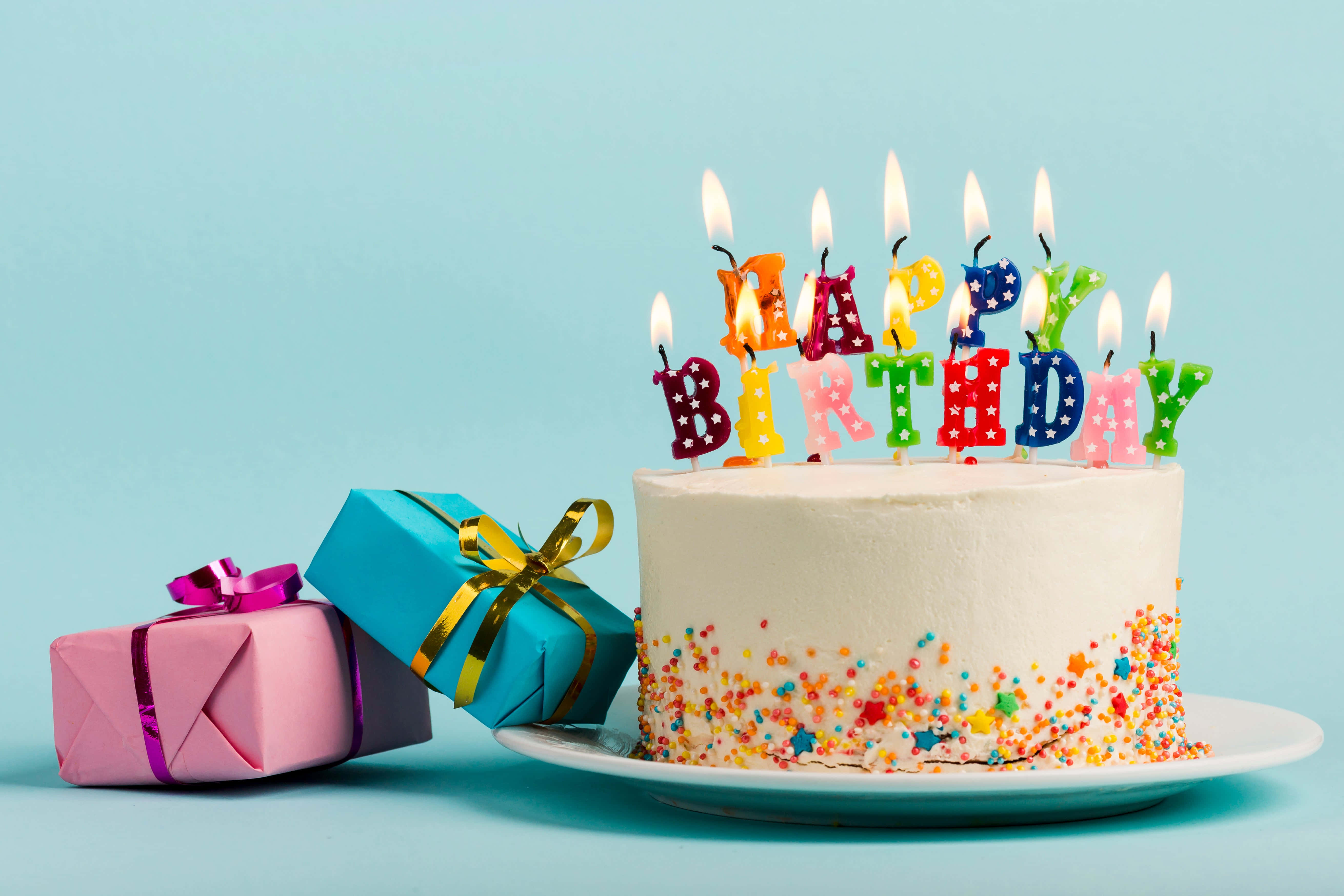 生日蛋糕 桌布 壁紙 — 一個白色生日蛋糕，上面有彩色字母蠟燭拼出 'HAPPY BIRTHDAY' 和星星糖粒，旁邊有兩個包裝好的禮物，放在淺藍色背景上。