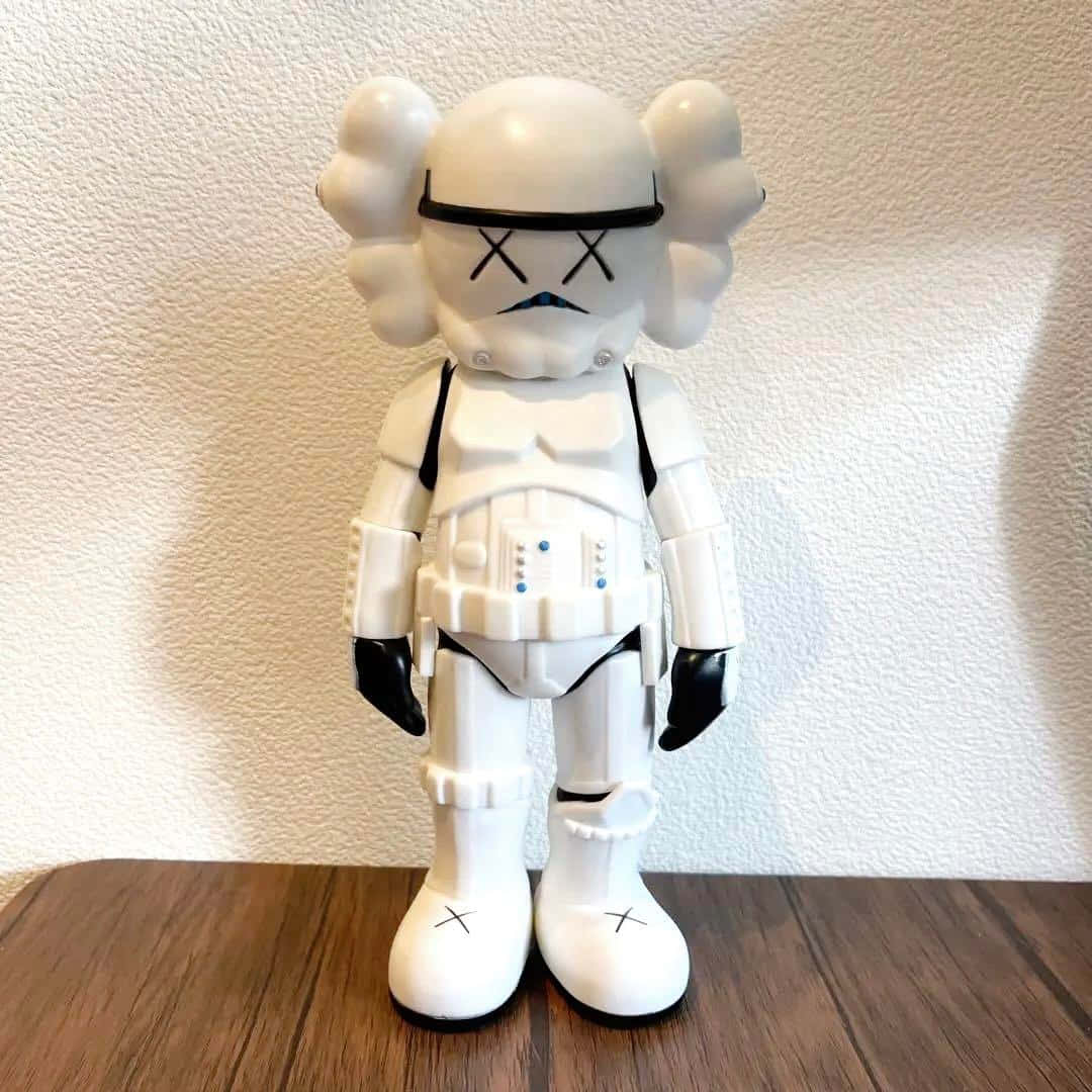 Kaws Stormtrooper 桌布 壁紙 — 一個白色的KAWS風暴兵公仔，帶有X形眼睛和雲朵狀耳朵，站在木質表面上，背景是紋理牆壁。