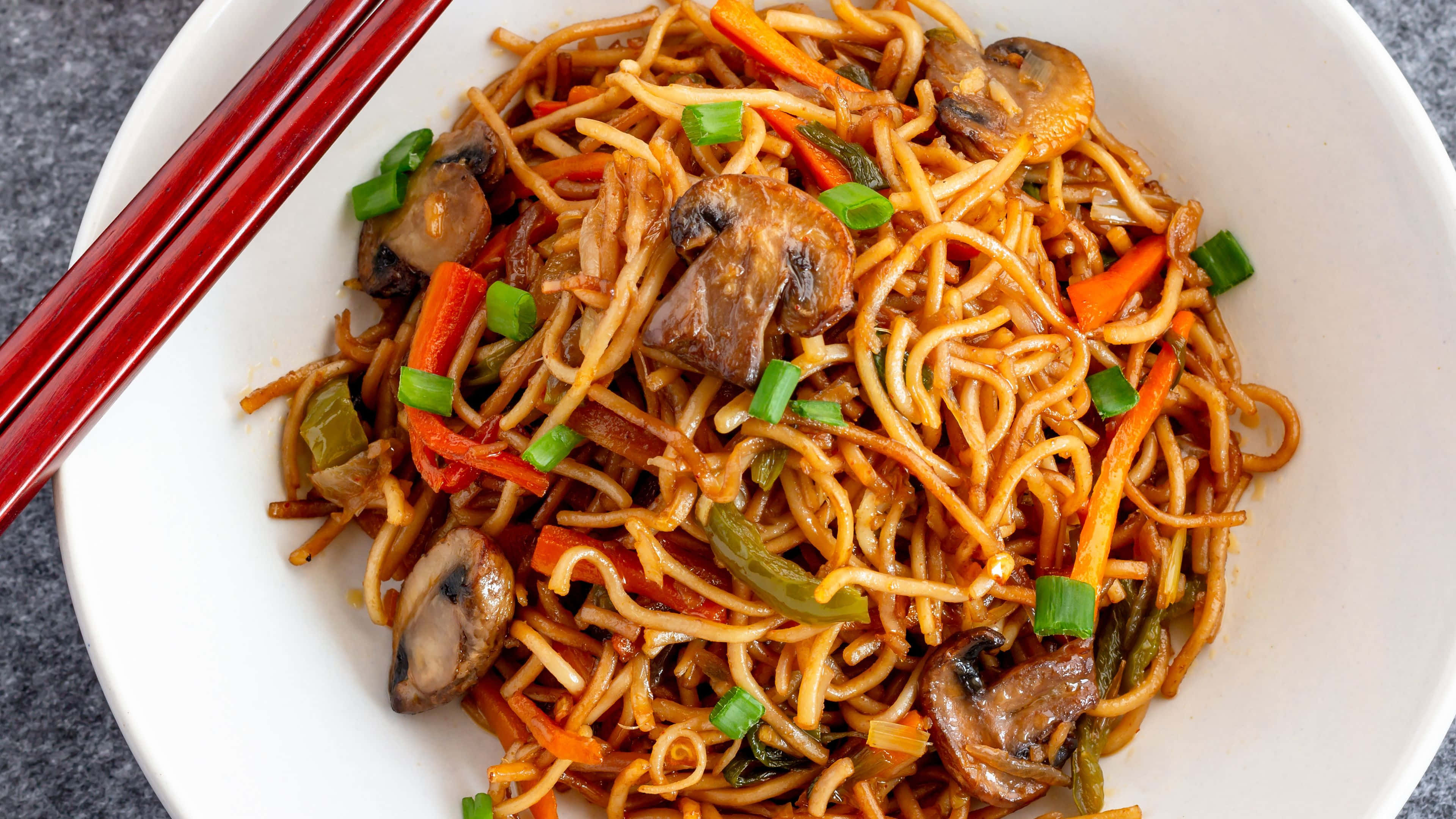 Noodles 桌布 壁紙 — 一個白色碗中的炒麵，配有蘑菇、紅蘿蔔和青蔥，並有紅色筷子，特寫鏡頭。