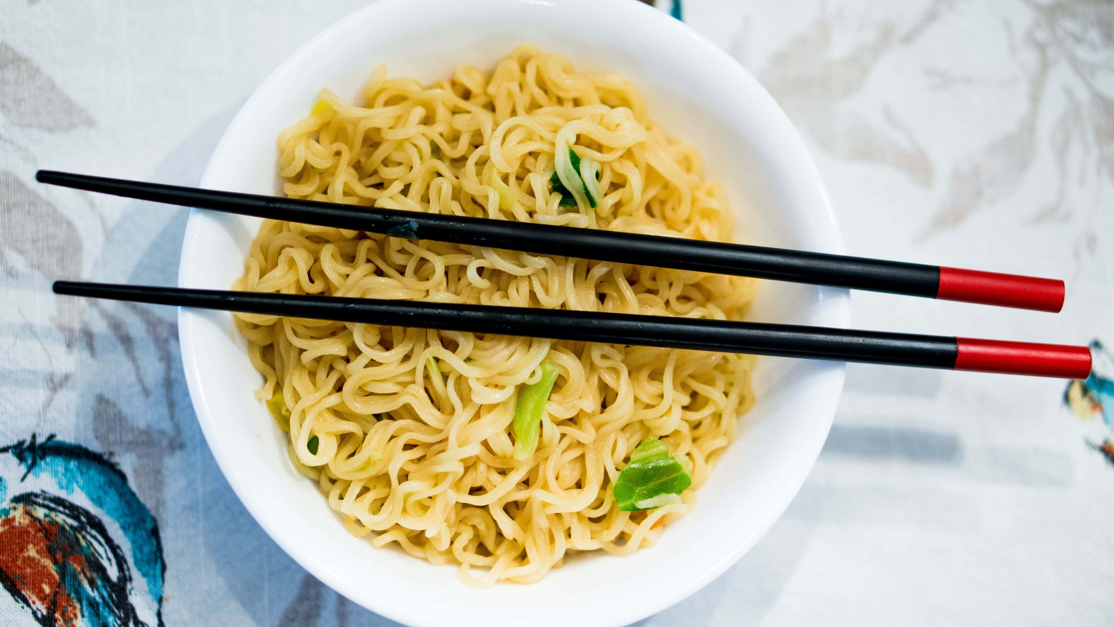 Noodles 桌布 壁紙 — 一個白色碗的俯視圖，裡面裝滿了冒著熱氣的拉麵，上面裝飾著青蔥，並放著一對尖端紅色的黑色筷子。