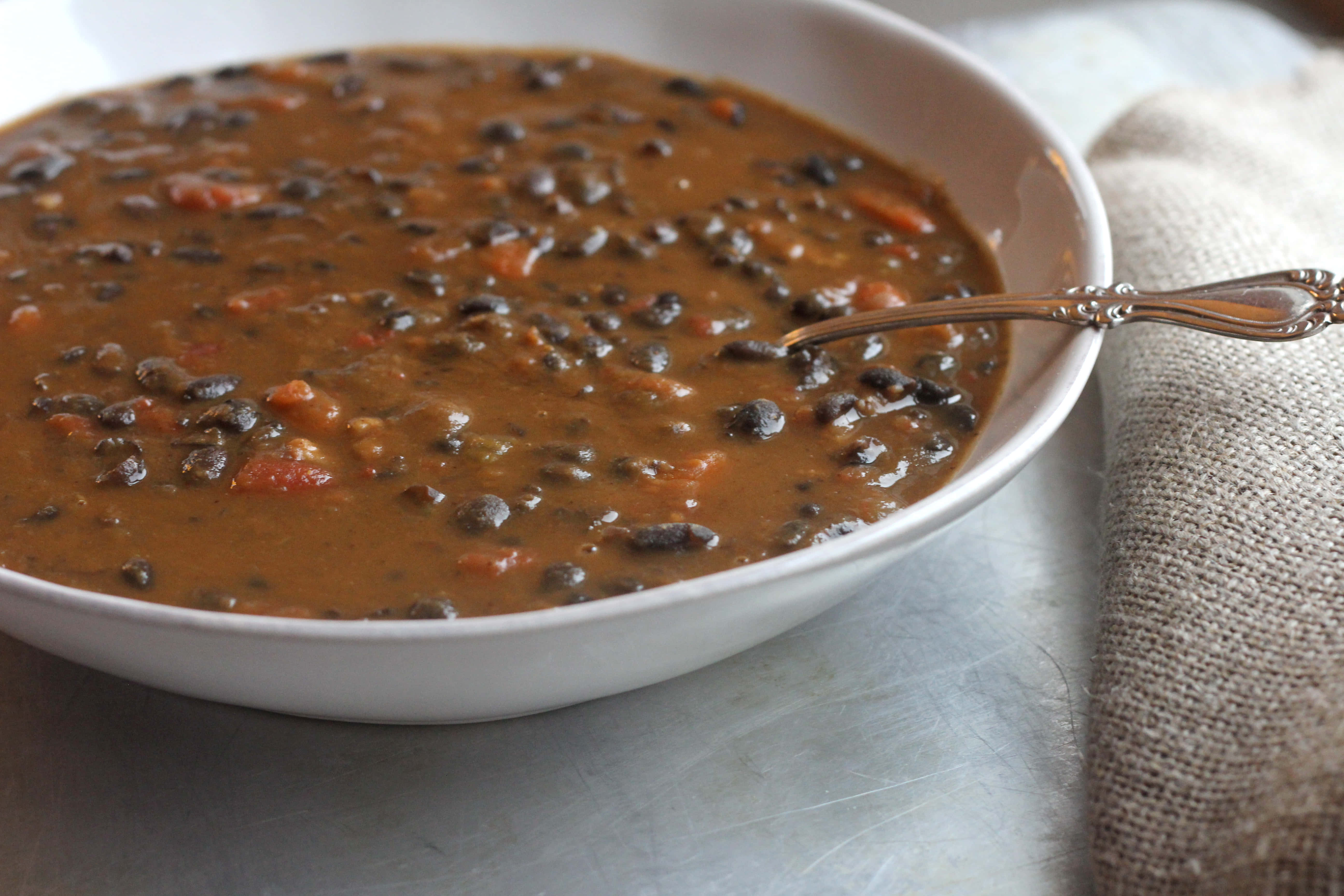 Black Bean Soup 桌布 壁紙 — 一個白色碗的特寫，裡面裝滿了濃郁的棕色黑豆湯，可見番茄塊，並有一隻復古湯匙放在裡面。