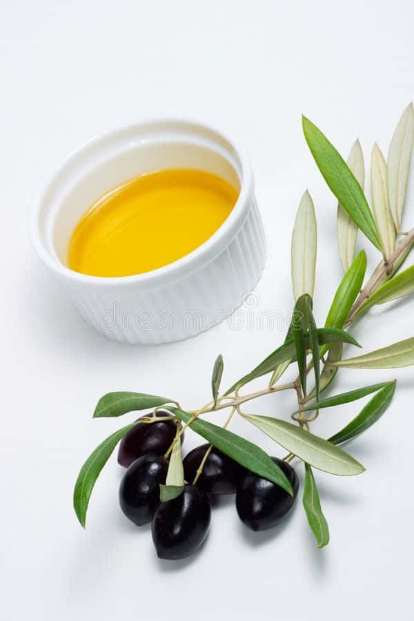 Black Olives 桌布 壁紙 — 一個白色碗盛著金黃色的橄欖油，旁邊放著一枝帶有深色橄欖和綠葉的樹枝，放在潔白的表面上。