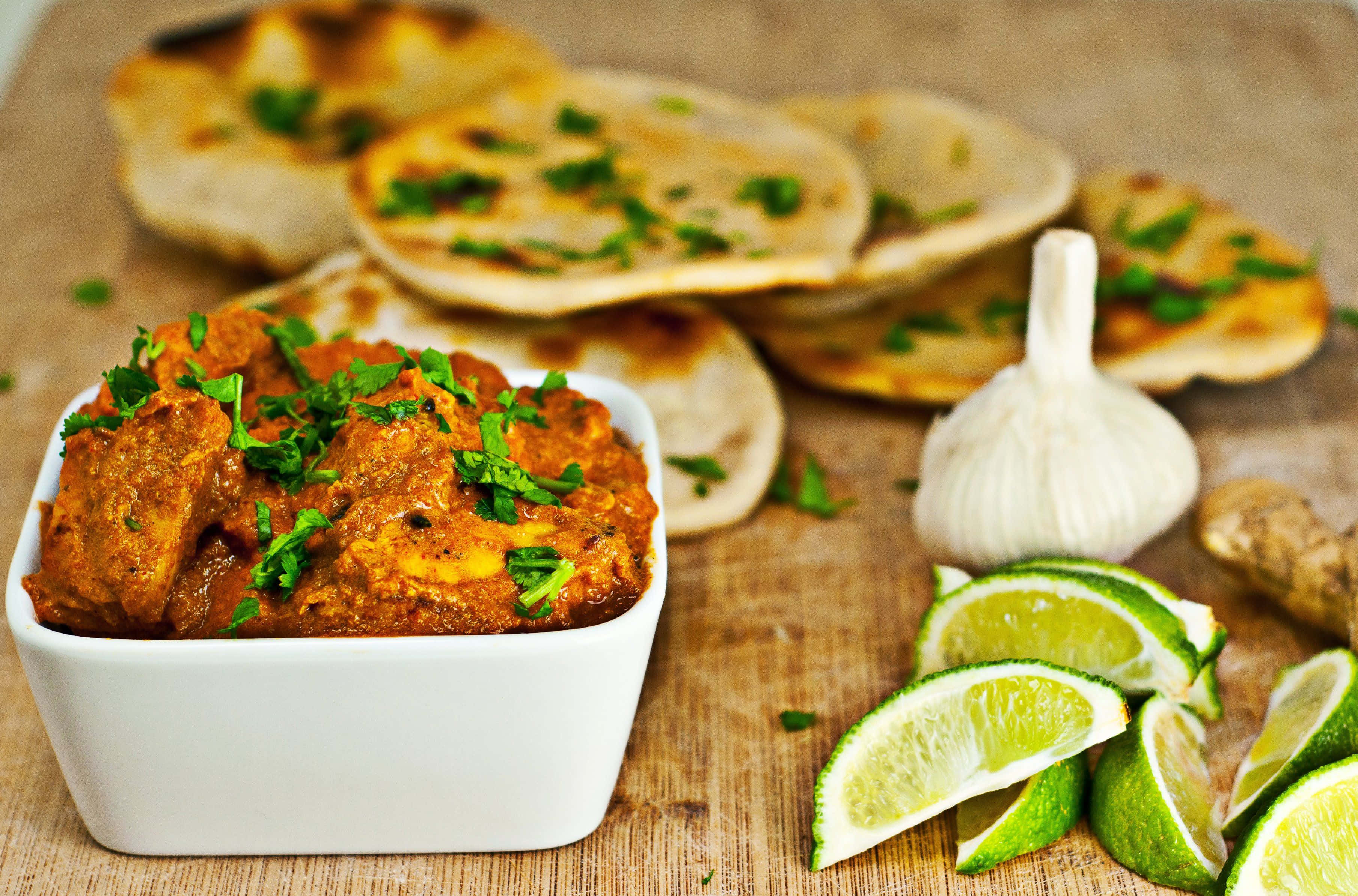 Indian Food 桌布 壁紙 — 一個白色碗盛載著濃郁的橙色咖喱，配上芫荽裝飾，旁邊有青檸角、蒜頭和烤餅放在木板上。