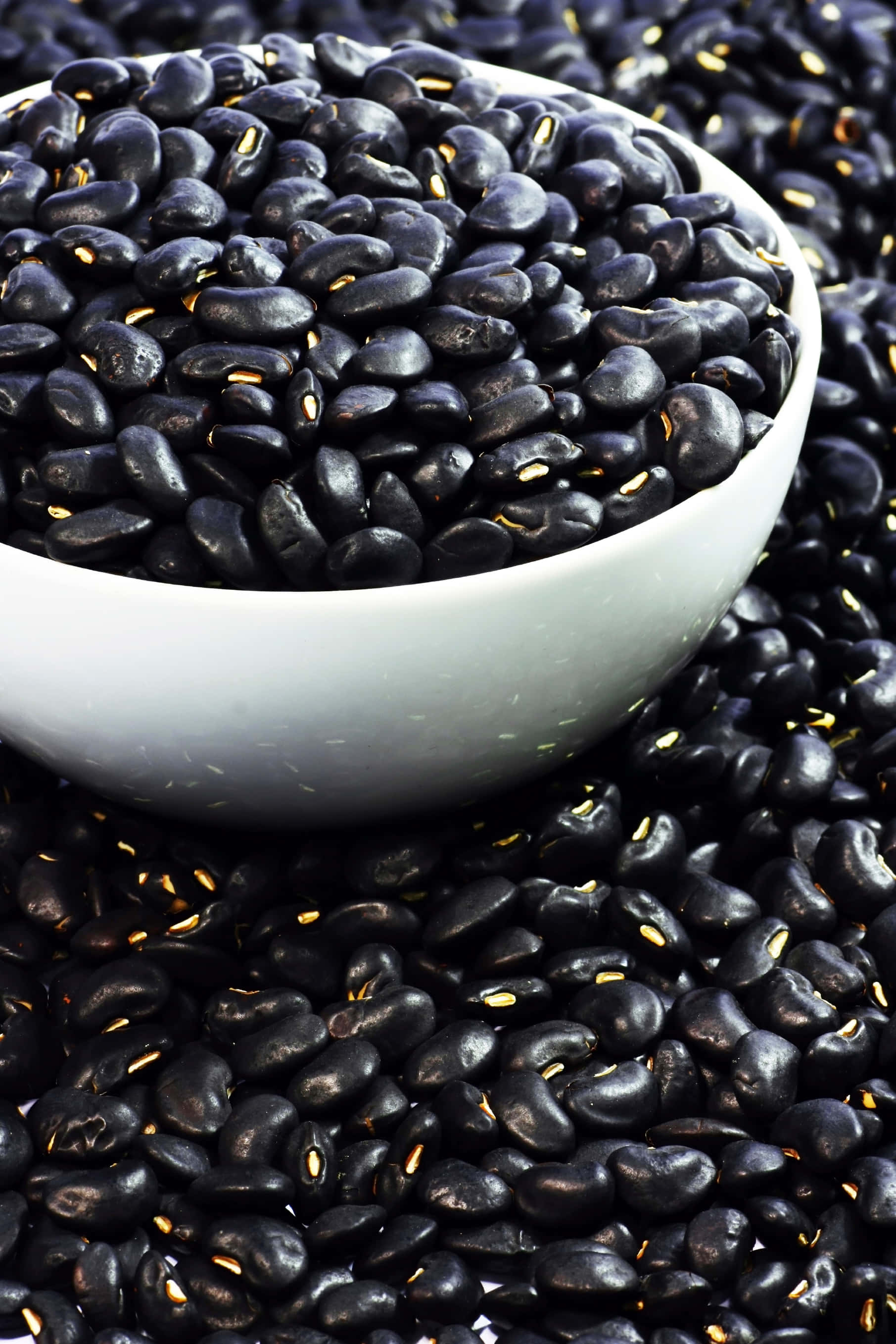 beans 桌布 壁紙 — 一個白色碗裝滿了光澤的黑豆，周圍還有更多豆子，在明亮光線下露出金色種臍。