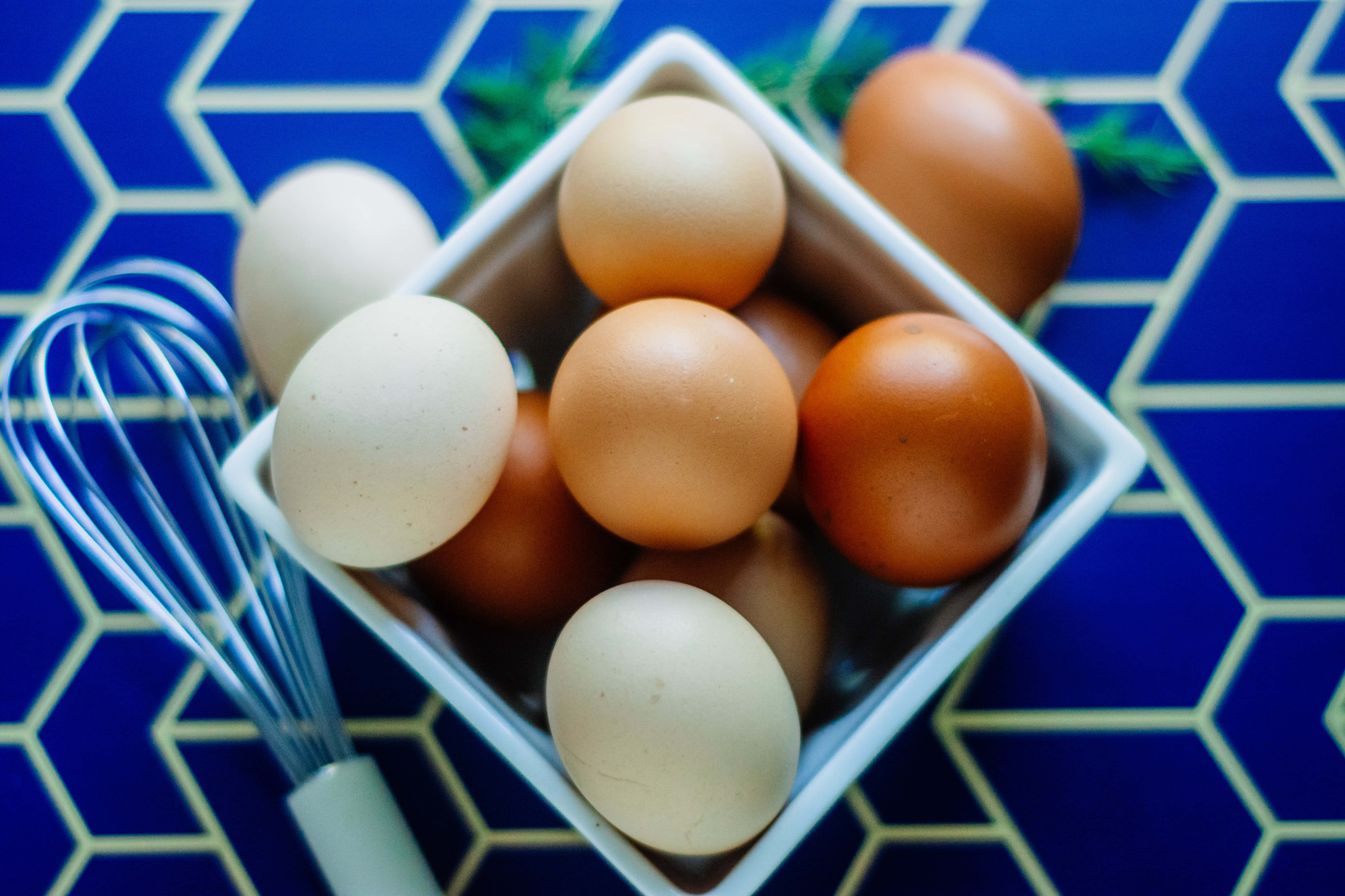 Brown Eggs 桌布 壁紙 — 一個白色碗裝滿棕色和奶油色的雞蛋，放在藍色六角形瓷磚表面上，旁邊有一個金屬打蛋器。
