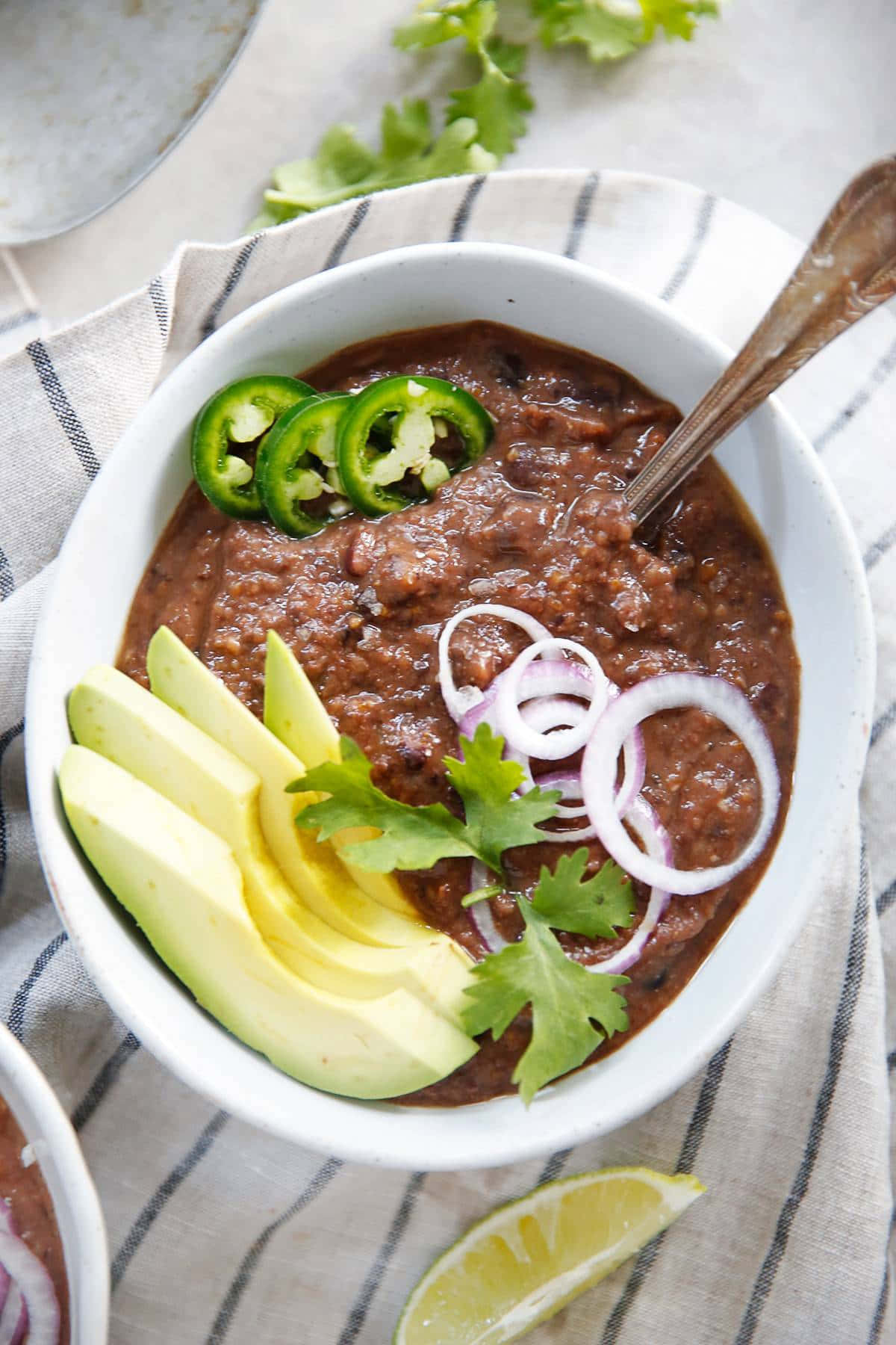 Black Bean Soup 桌布 壁紙 — 一個白色碗裝滿濃郁的深色辣椒，上面放有牛油果片、紅洋蔥圈、墨西哥辣椒片和新鮮芫荽。