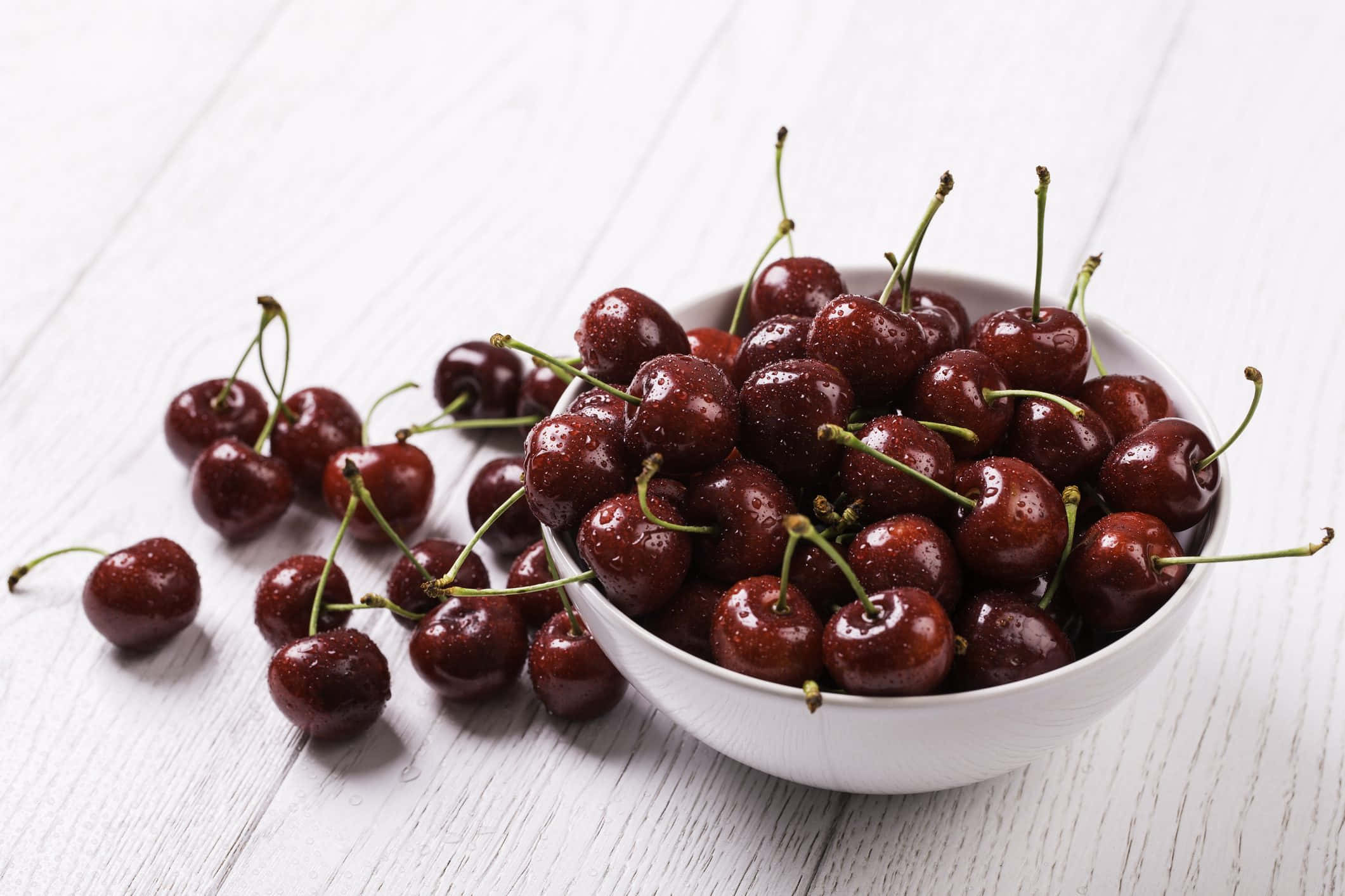 Black Cherry 桌布 壁紙 — 一個白色碗裝滿閃亮的深紅色櫻桃，放在磨損的白色木質表面上，附近散落著水果。