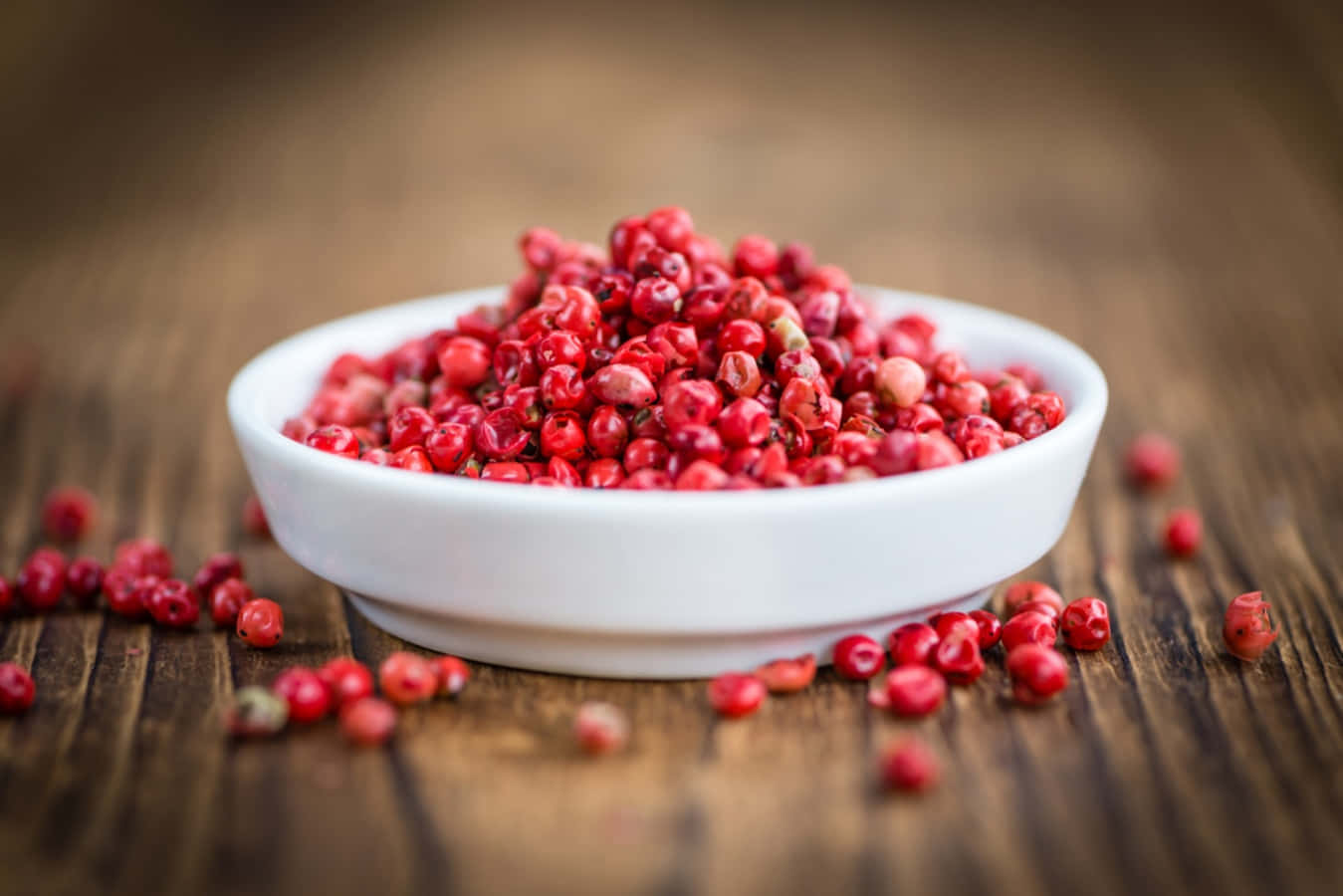 Pink Peppercorn 桌布 壁紙 — 一個白色碗裝滿鮮紅色胡椒粒，放在質樸的木質表面上，周圍散落著漿果。