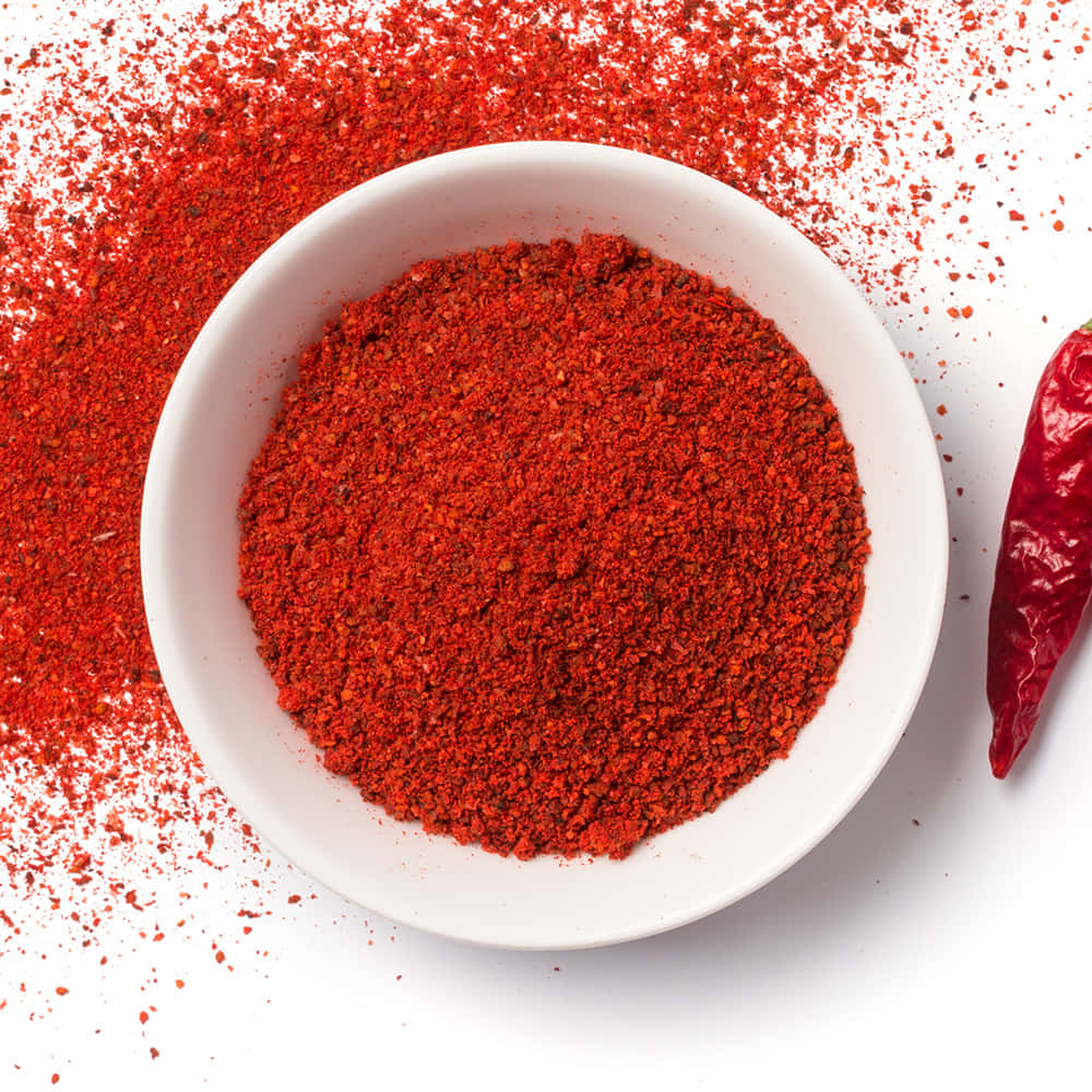 Red Chili Powder 桌布 壁紙 — 一個白色碗裝滿鮮紅色辣椒粉，周圍散落著香料，旁邊還有一整條乾紅辣椒，放在乾淨的白色表面上。