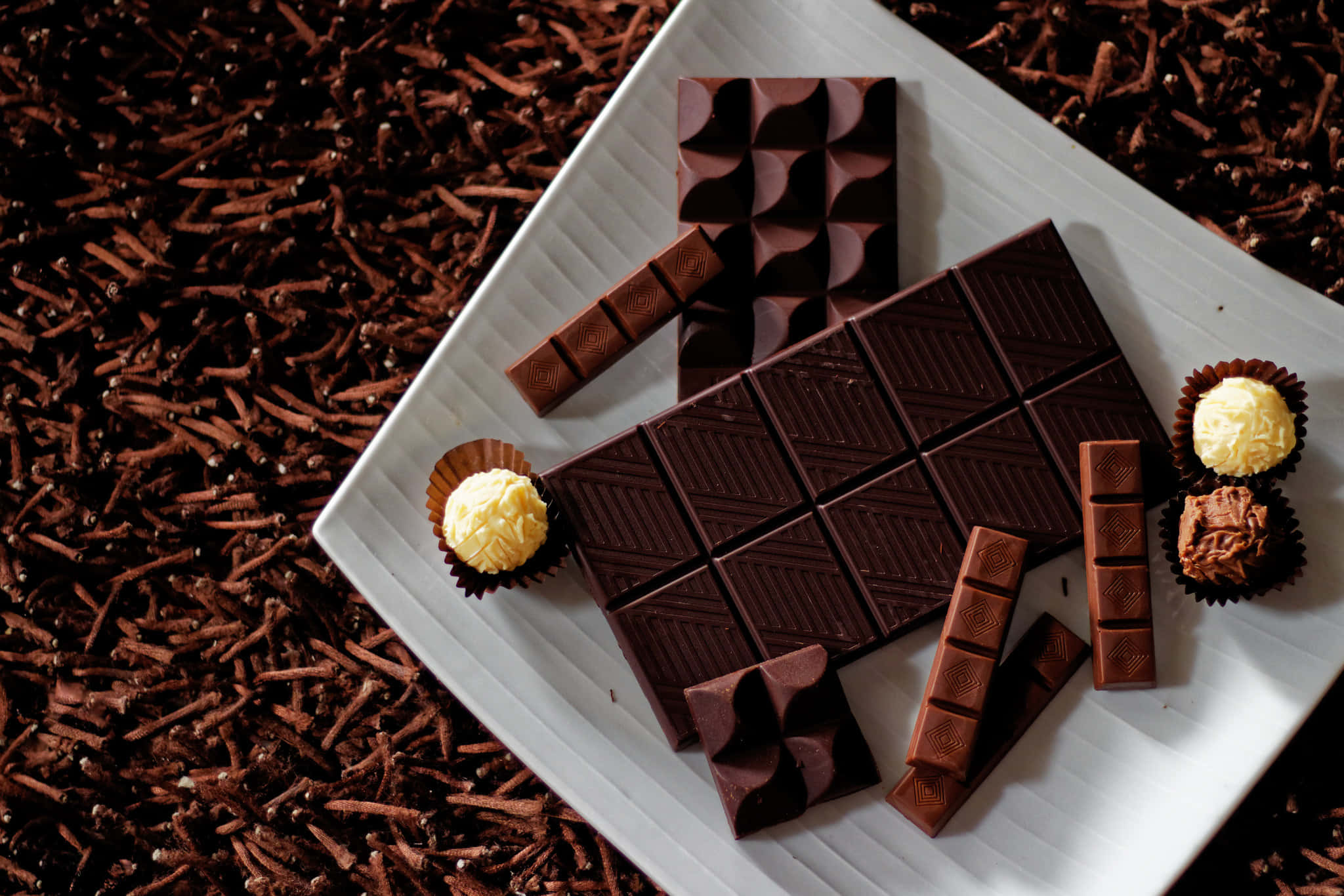 Dark Chocolate 桌布 壁紙 — 一個白色碟子上放著各式各樣的黑朱古力條和松露朱古力，周圍是可可碎粒，背景是豐富的棕色。