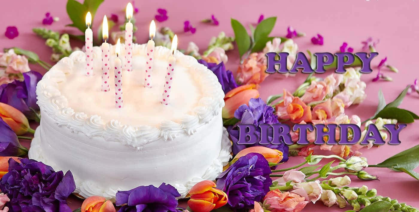 Birthday Wishes 桌布 壁紙 — 一個白色糖霜生日蛋糕，上面插著點燃的蠟燭，周圍環繞著鮮豔的紫色和橙色花朵，背景為粉紅色。