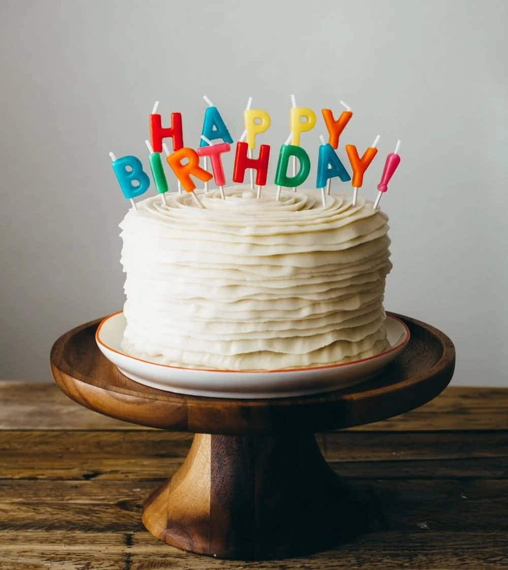 生日蛋糕 桌布 壁紙 — 一個白色糖霜生日蛋糕，上面有彩色字母蠟燭拼出 happy birthday，放在木製蛋糕架上。