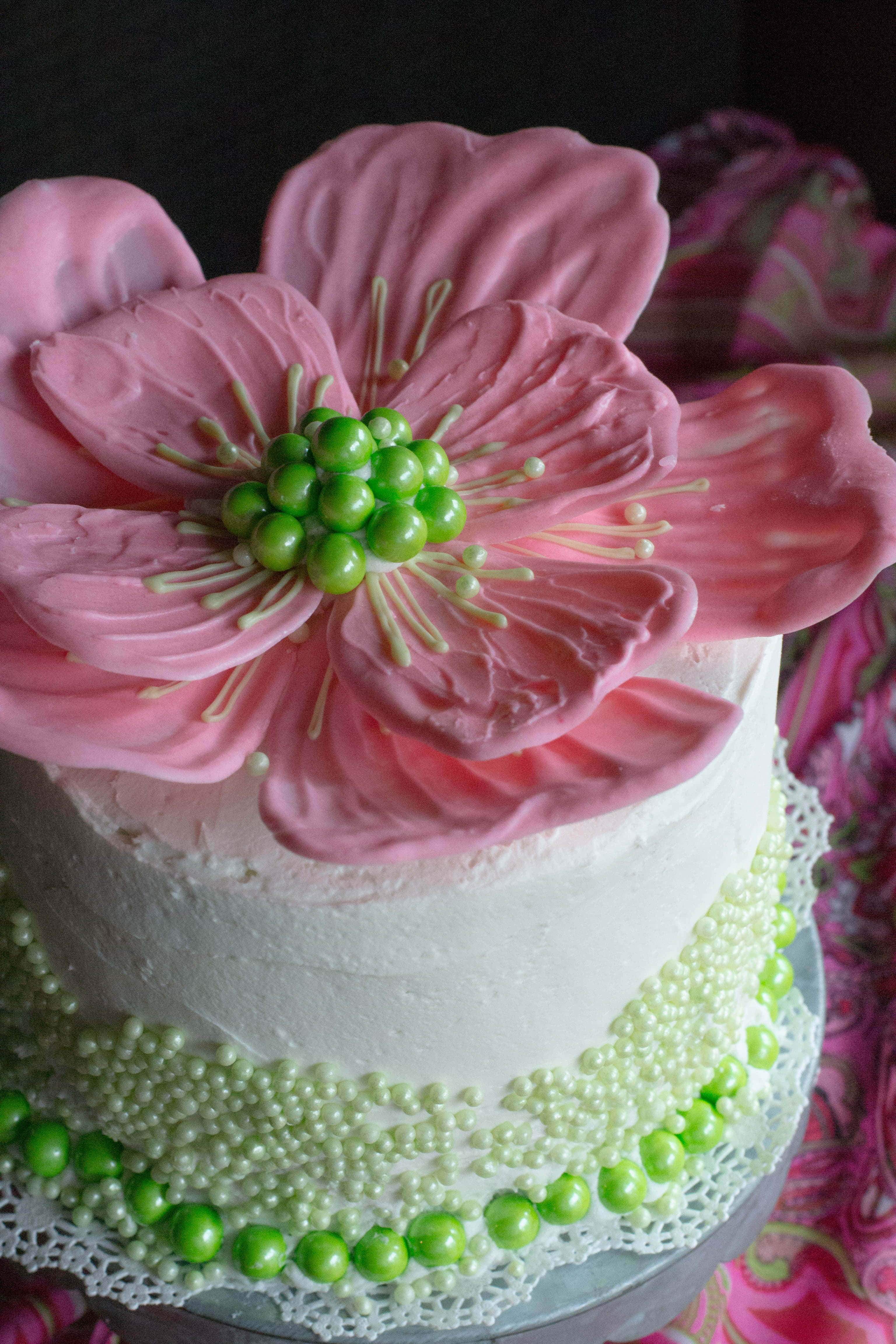 Floral Cake 桌布 壁紙 — 一個白色糖霜蛋糕，上面裝飾著一朵大的粉紅色糖花和綠色珠子點綴，放在蕾絲墊上。