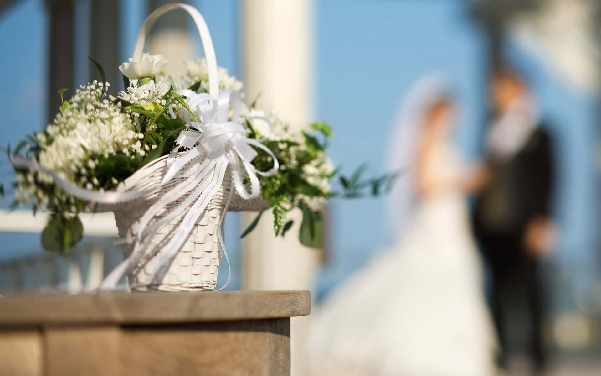 Wedding Bouquet 桌布 壁紙 — 一個白色編織籃子，裝滿精緻的白色花朵和綠葉，在木質表面上清晰對焦，背景中有一對模糊的新娘和新郎站在明亮的藍天下。