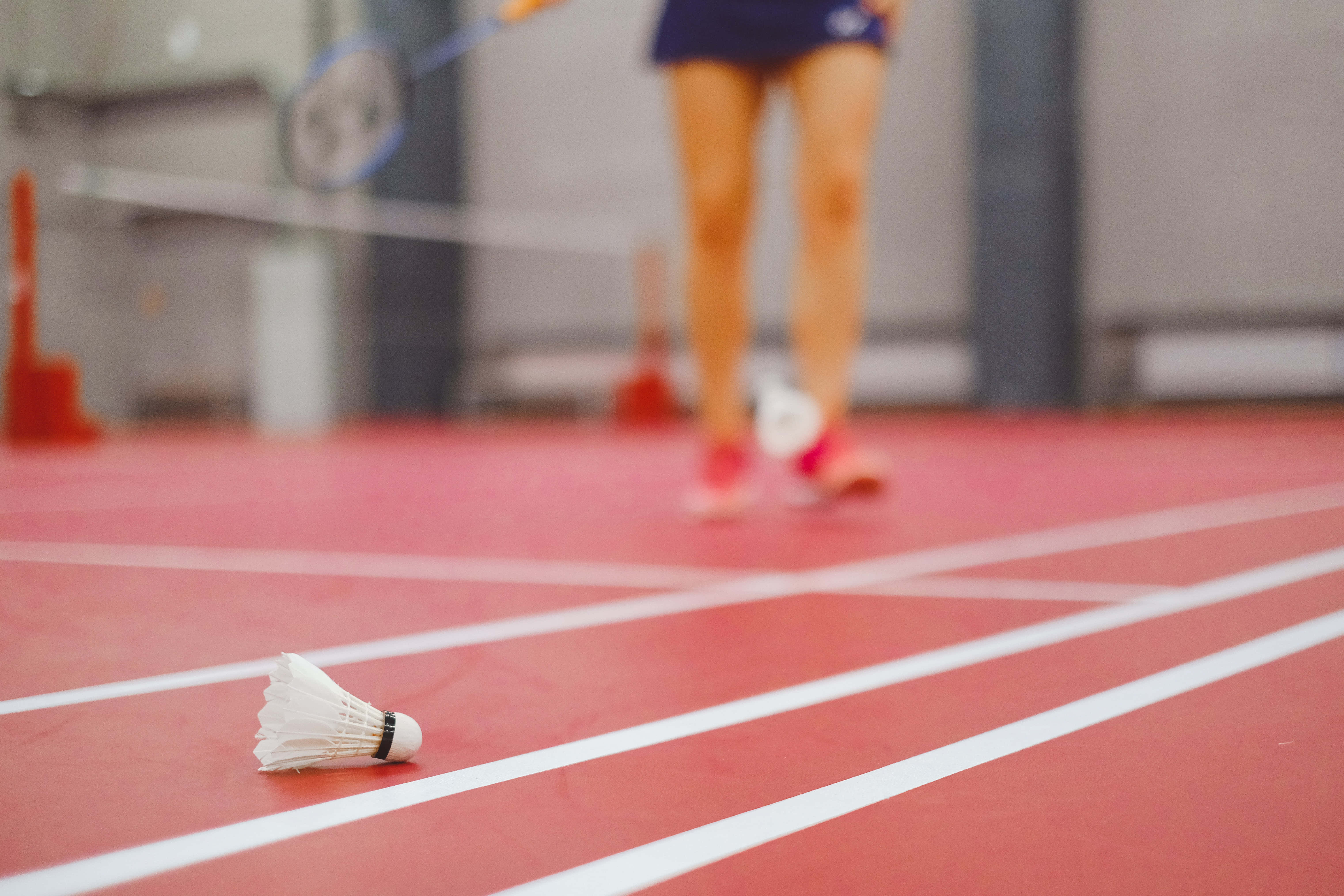 Badminton 桌布 壁紙 — 一個白色羽毛球放在紅色羽毛球場上，背景中移動的球員變得模糊。