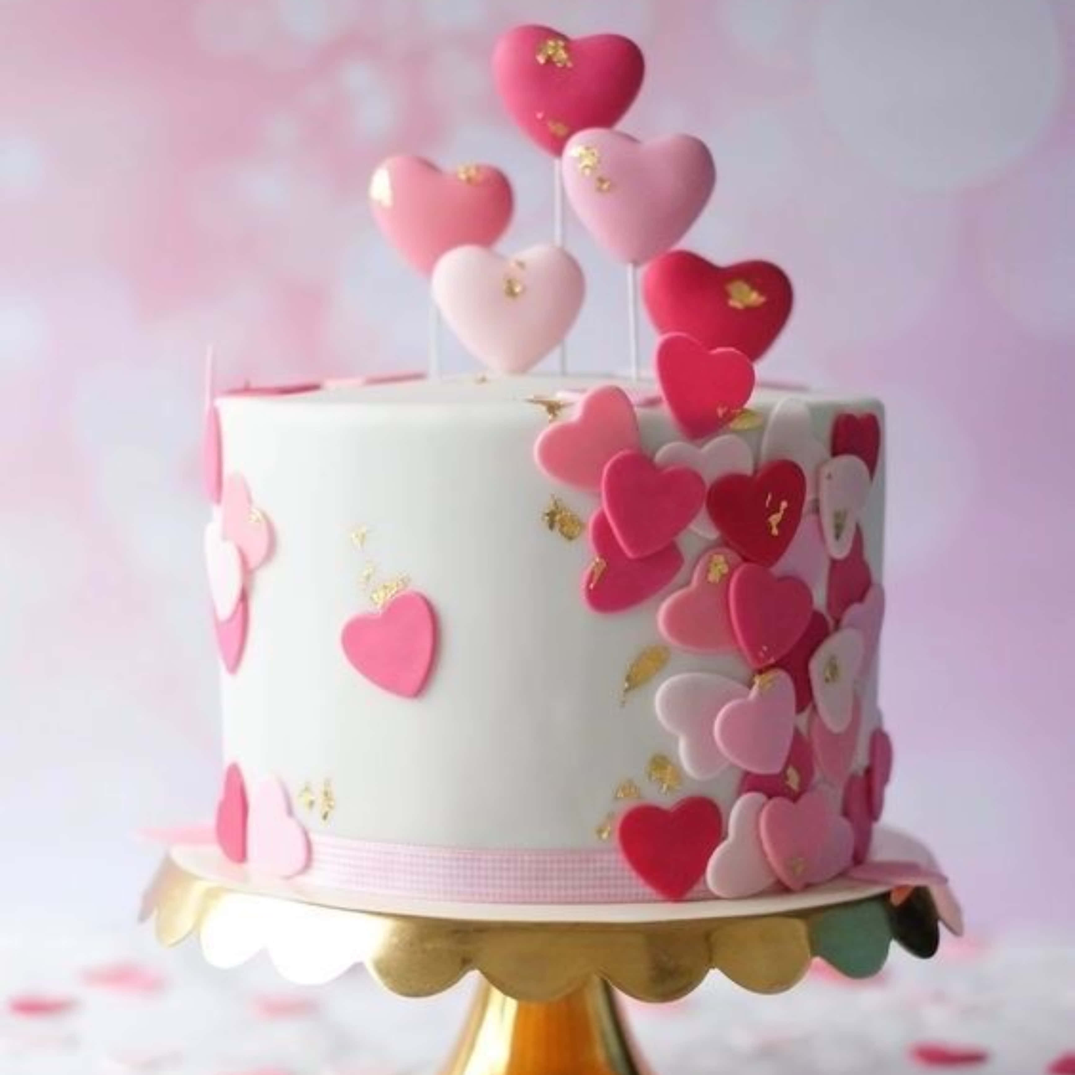 Cake Fondant 桌布 壁紙 — 一個白色蛋糕，飾有粉紅色和紅色的心形及金箔，放在金色架子上，背景是柔和的粉紅色。