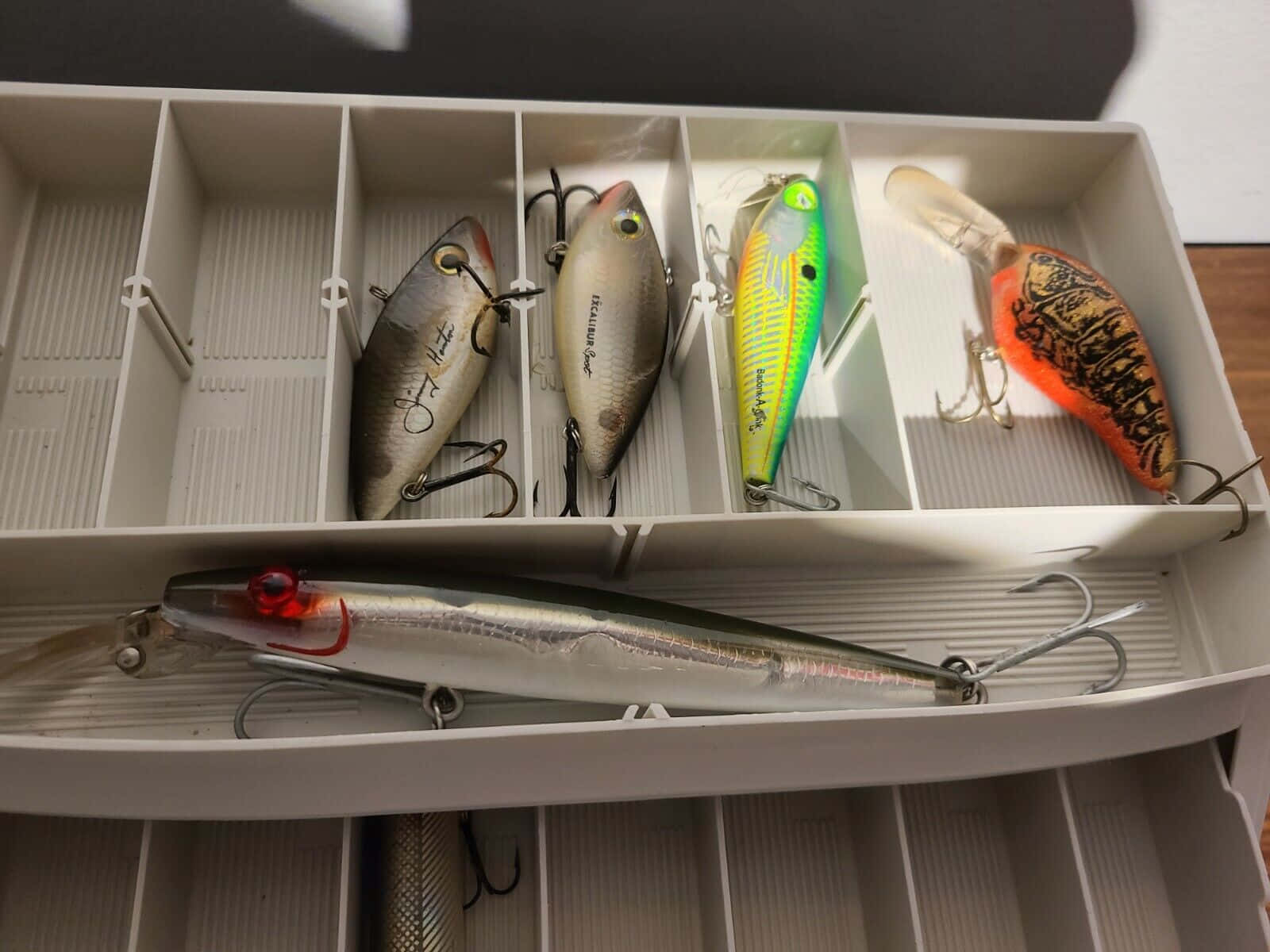 Fishing Bait 桌布 壁紙 — 一個白色釣具箱內有多個隔層，裝有各種魚餌，前面放著一個大銀色魚餌。