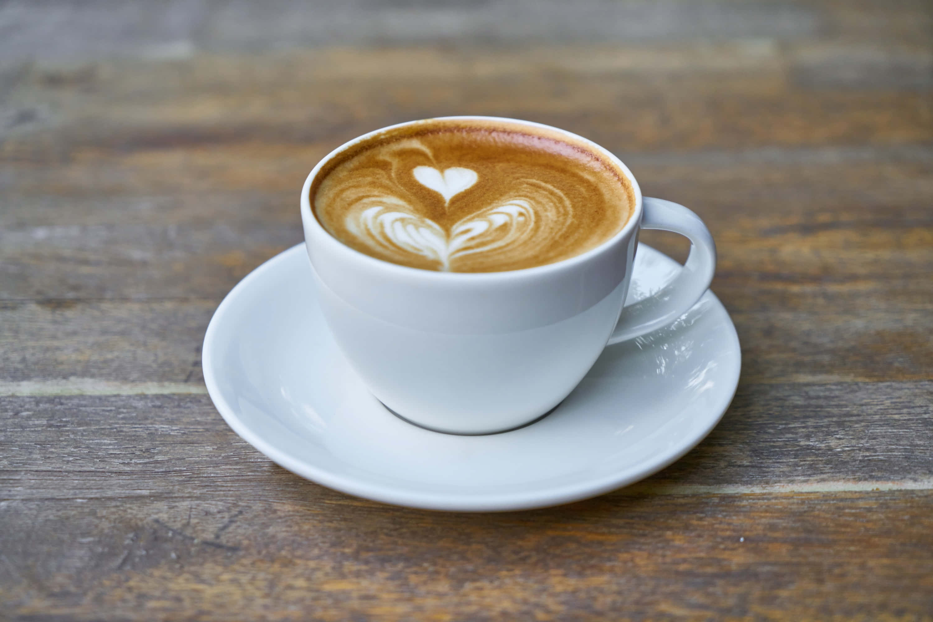 Coffee Cup 桌布 壁紙 — 一個白色陶瓷杯，上面有心形拉花藝術，放在一個樸實的木桌上嘅碟子上。