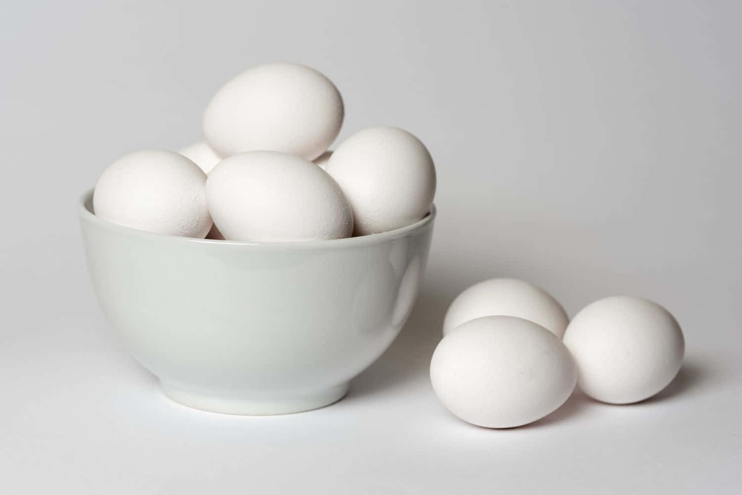 Egg 桌布 壁紙 — 一個白色陶瓷碗裝滿光滑的白雞蛋，旁邊有三個散落的雞蛋，放在極簡的白色表面上。