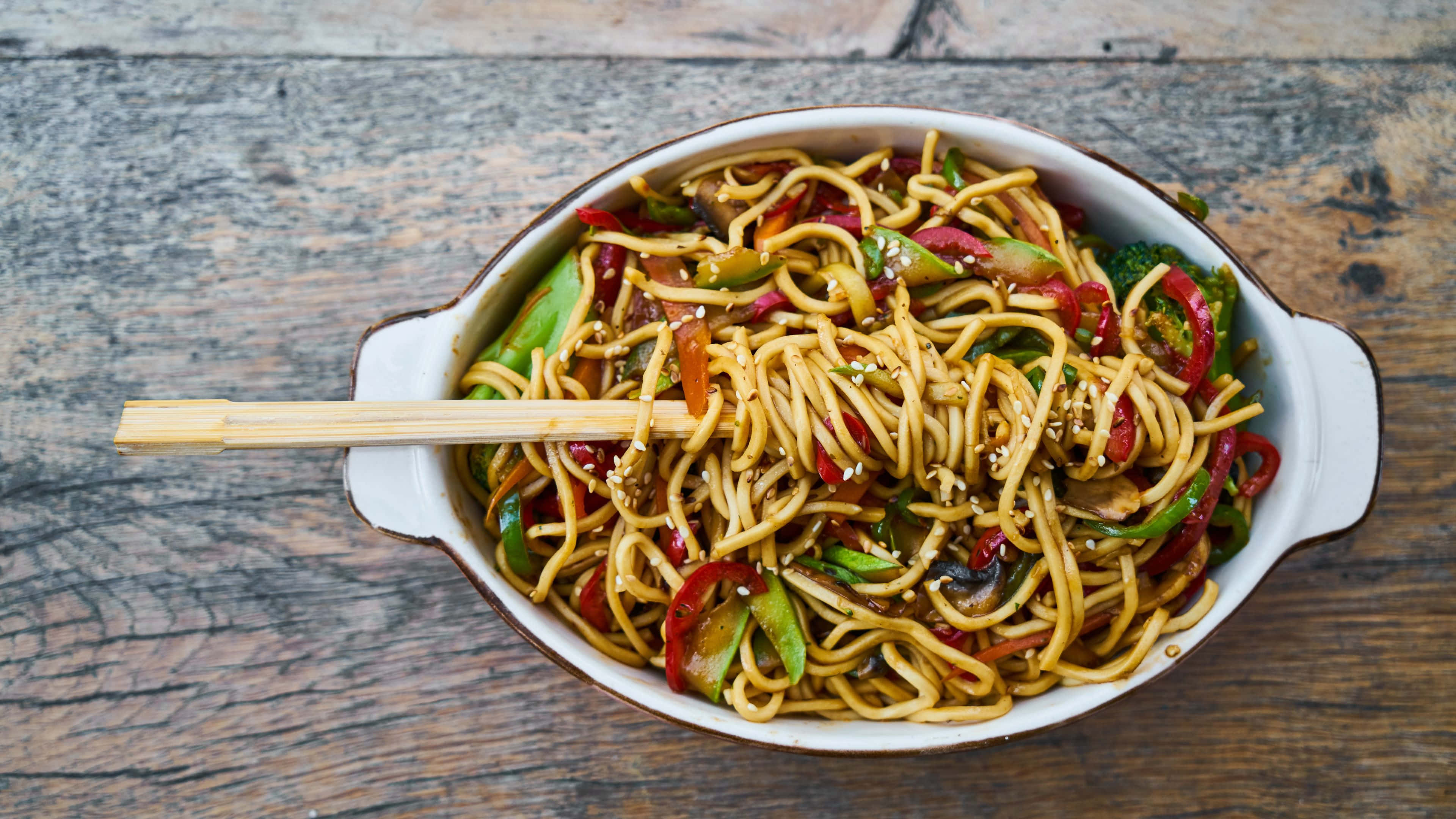 Noodles 桌布 壁紙 — 一個白色陶瓷碗裝滿炒麵和色彩繽紛的蔬菜，放在質樸的木桌上，筷子擱在旁邊。