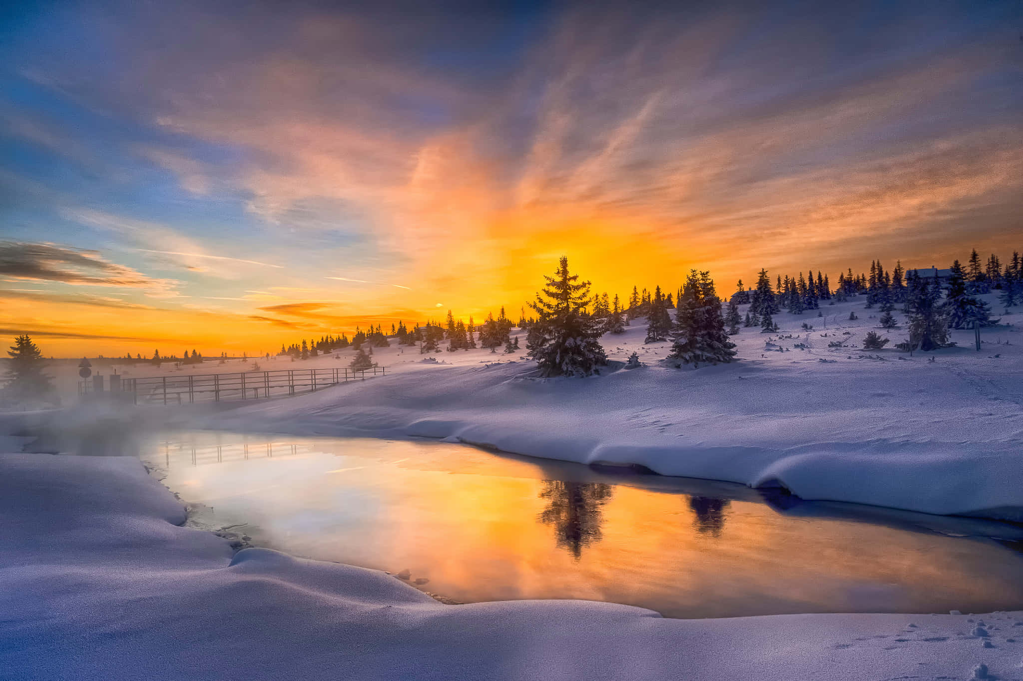 Winter Sunset 桌布 壁紙 — 一個白雪皚皚的冬季日出，將溫暖的金色光芒投射在一條霧氣瀰漫的河流上，河流映照著天空和積雪的松樹。