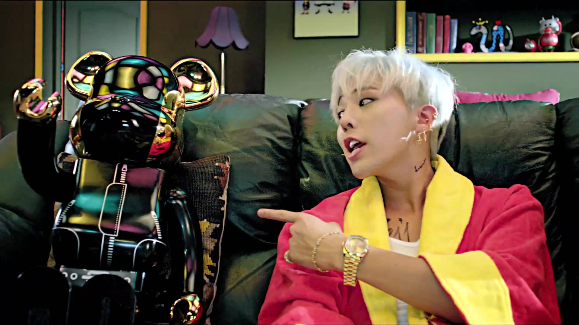 G-DRAGON《Crayon》 壁紙 — 一個白髮的人穿著紅色長袍，坐在深色沙發上指著一個閃亮的黑色熊雕像。