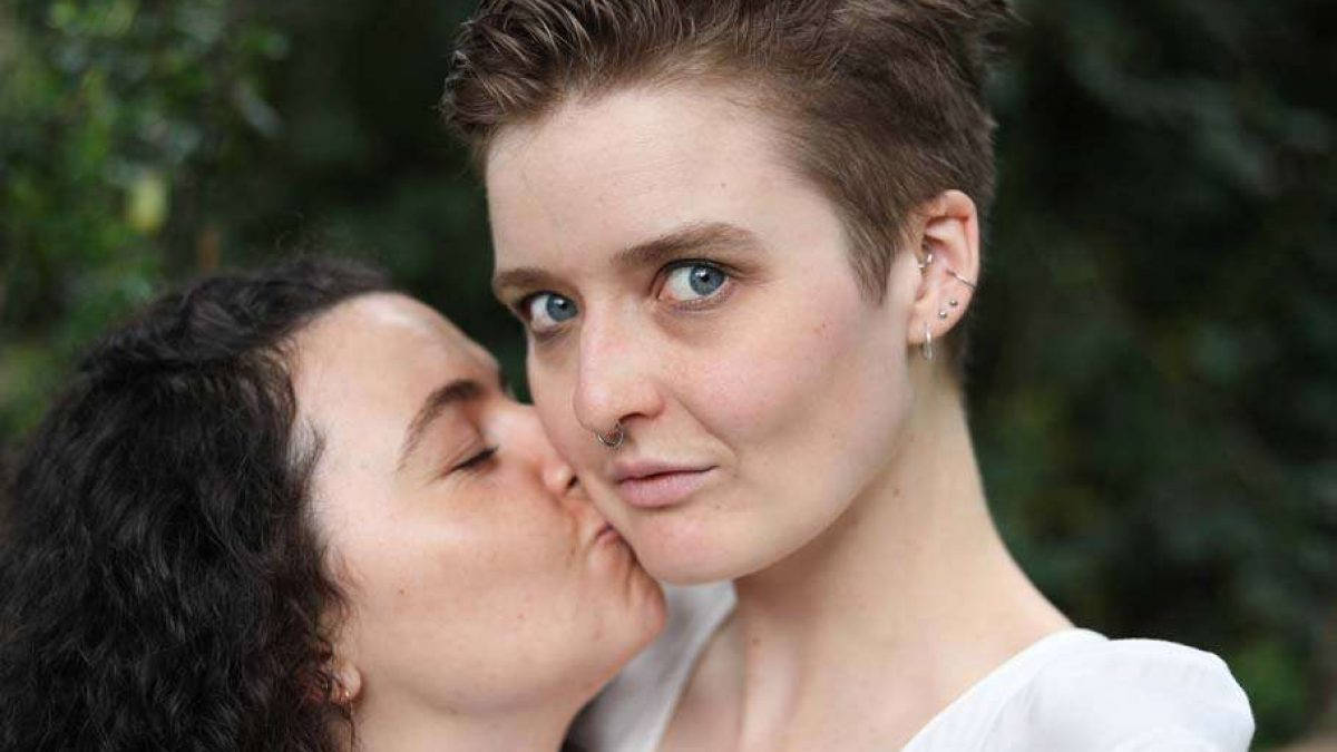 Two Girls Sharing a Tender Kiss 壁紙 — 一個短髮藍眼的人向前望，另一個人親吻他們的臉頰，在一個鬱鬱蔥蔥的綠色戶外環境中。