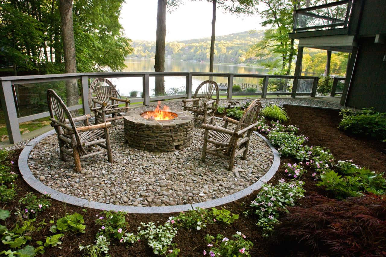 Garden Design 桌布 壁紙 — 一個石頭火坑，閃爍的火焰位於碎石圓圈內，周圍是質樸的木椅和盛開的花朵，位於一個現代化的甲板上，俯瞰著寧靜的湖泊。