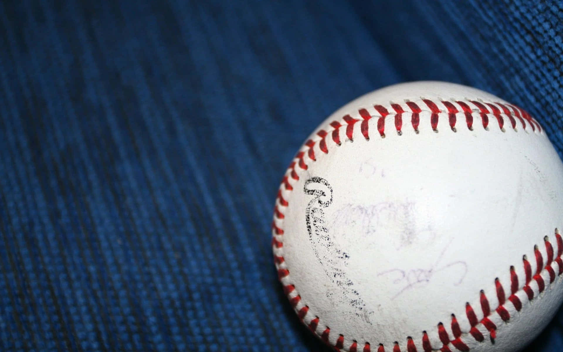 Cool Baseball 桌布 壁紙 — 一個磨損的棒球，帶有紅色縫線和褪色的簽名，放在有紋理的藍色布料上。