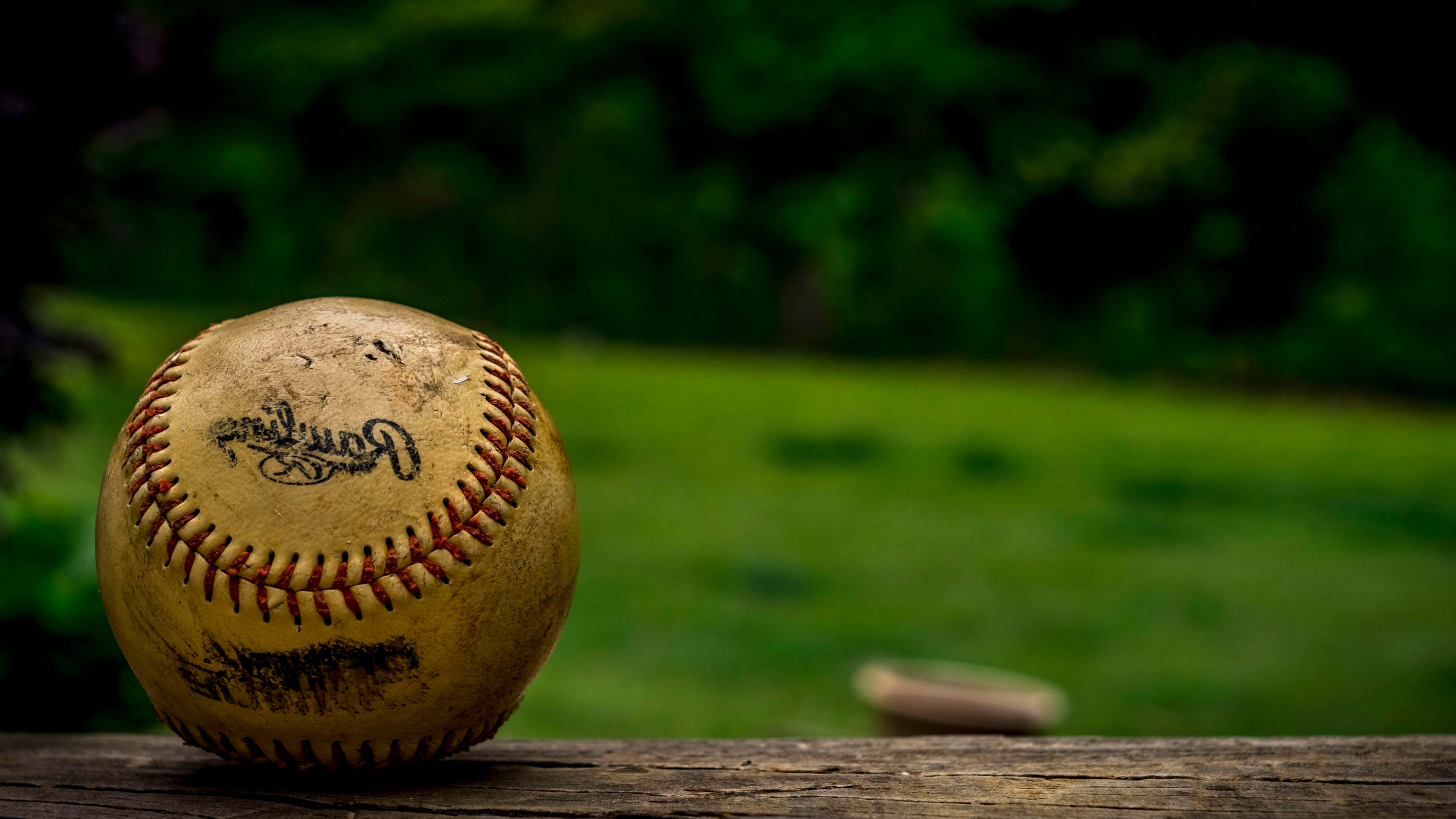 Cool Baseball 桌布 壁紙 — 一個磨損的棒球帶有紅色縫線，放在經風化的木質表面上，背景是模糊的綠色草地。