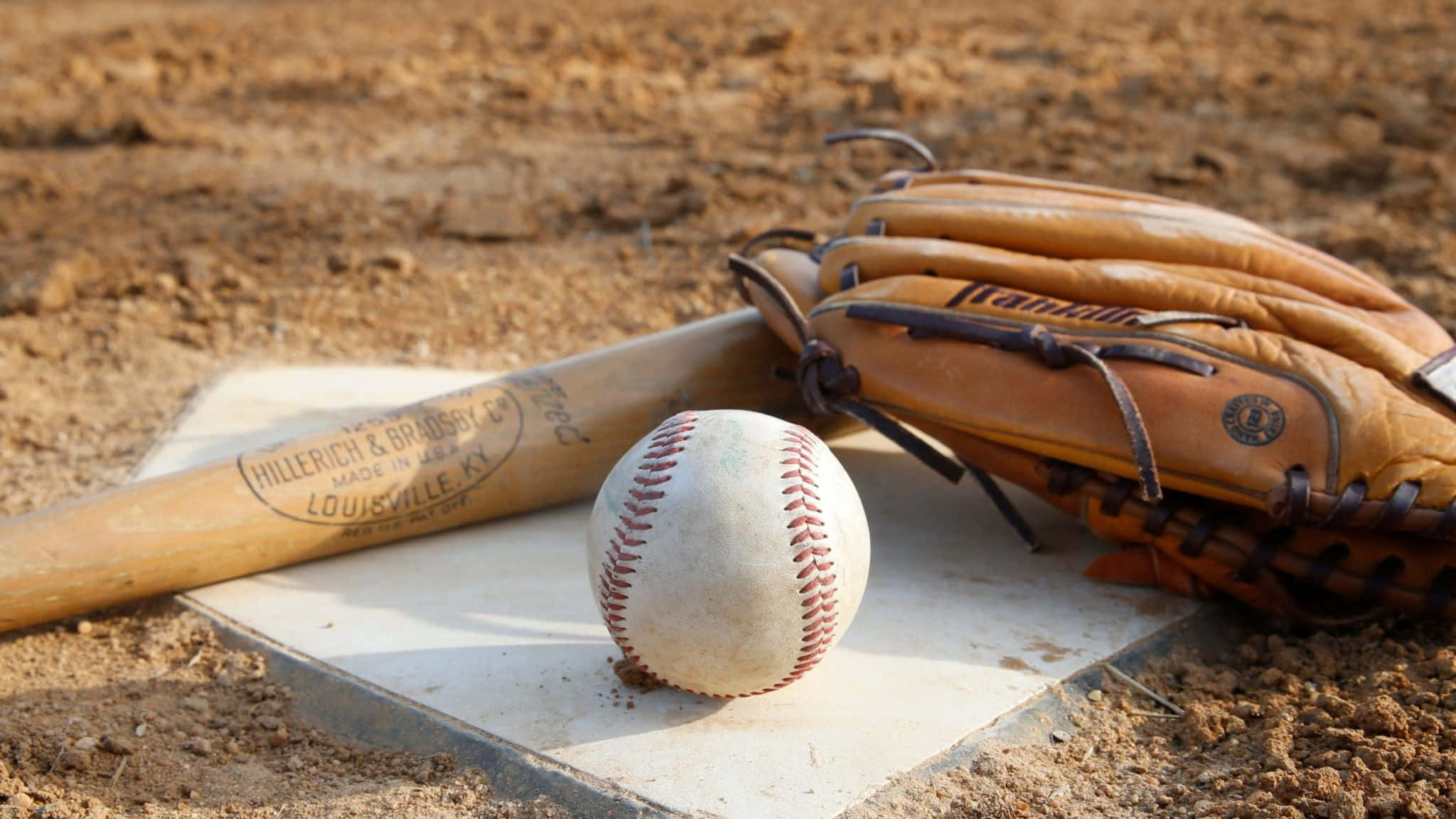 Baseball Gloves 桌布 壁紙 — 一個磨損的棒球棒、手套和球放在被泥土包圍的本壘板上，沐浴在溫暖的陽光下。