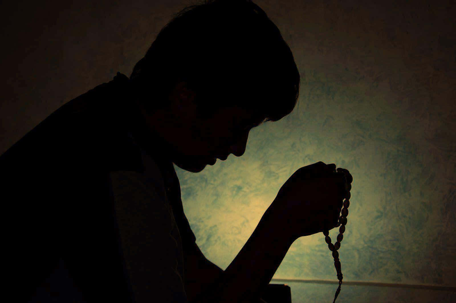 Praying 桌布 壁紙 — 一個祈禱的人嘅剪影，揸住玫瑰經念珠，靠住一面有紋理嘅牆，被溫暖嘅背光包圍。