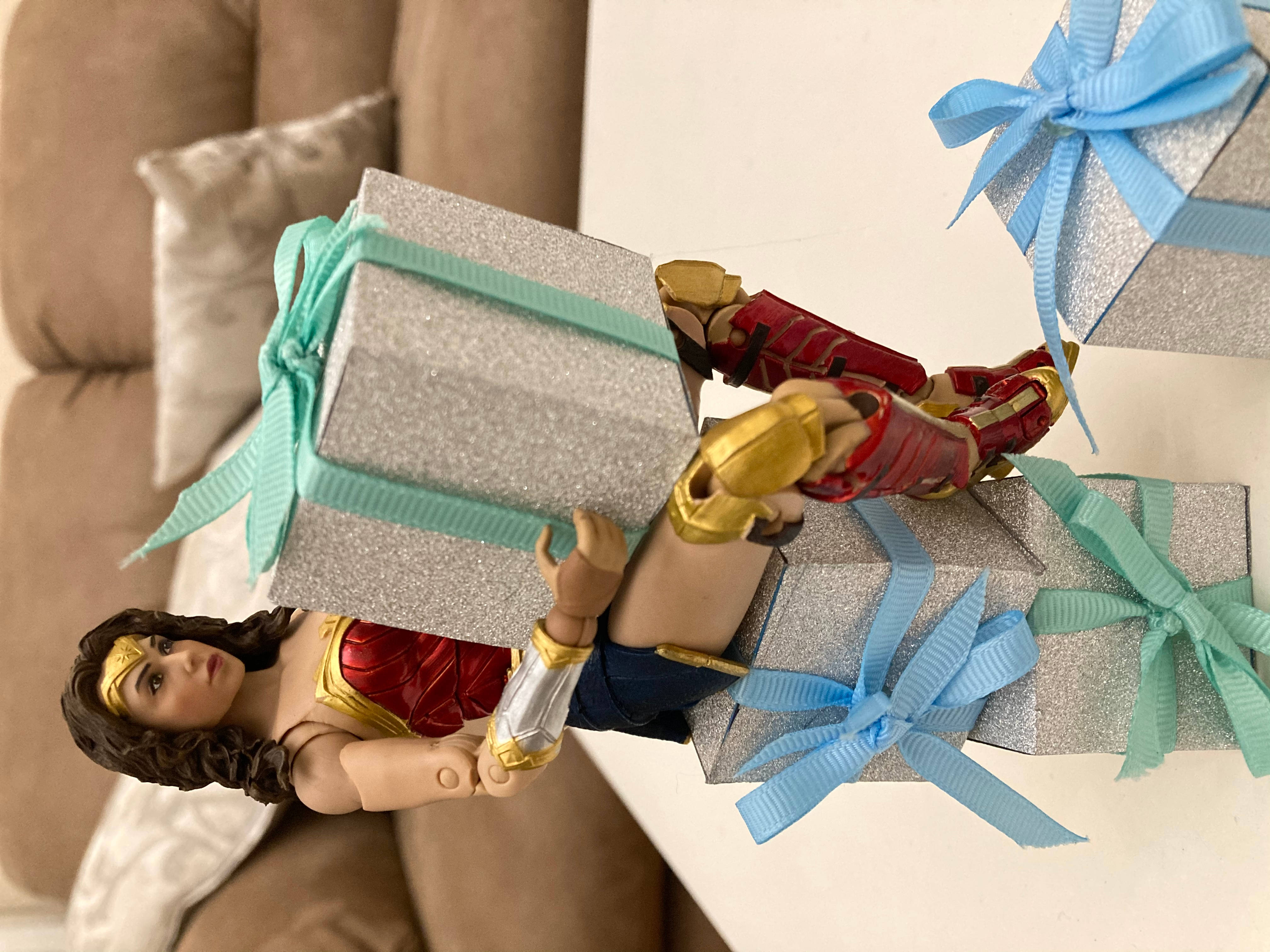 DC Comics Wonderwoman Toy 壁紙 — 一個神奇女俠動作人偶拿著閃閃發光的銀色禮物，上面有青色和藍色絲帶，背景是米色沙發。
