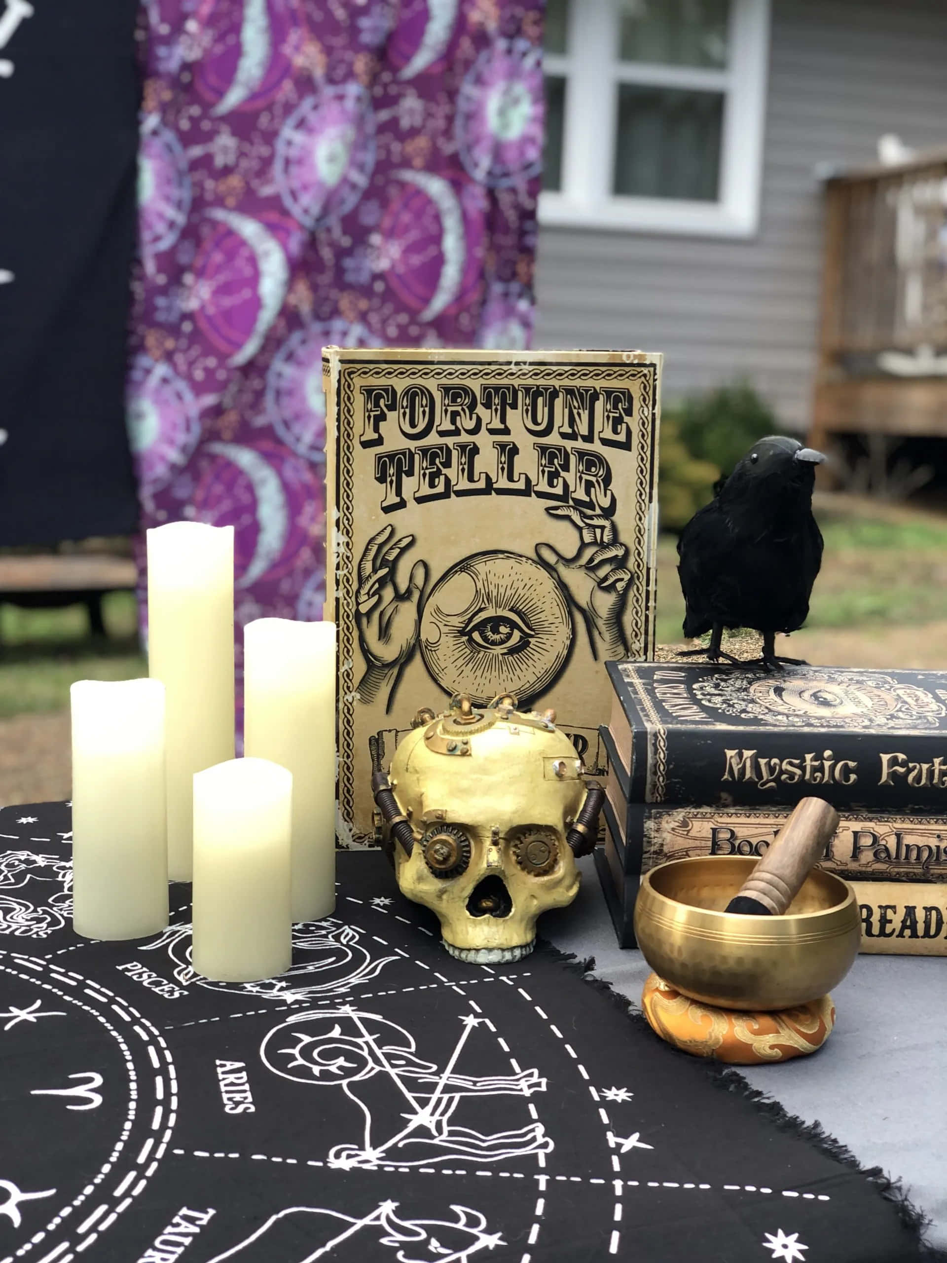 Fortune Teller 桌布 壁紙 — 一個神秘的戶外祭壇，有金色骷髏燭台、占卜書和一隻黑烏鴉棲息在神秘的書本上。